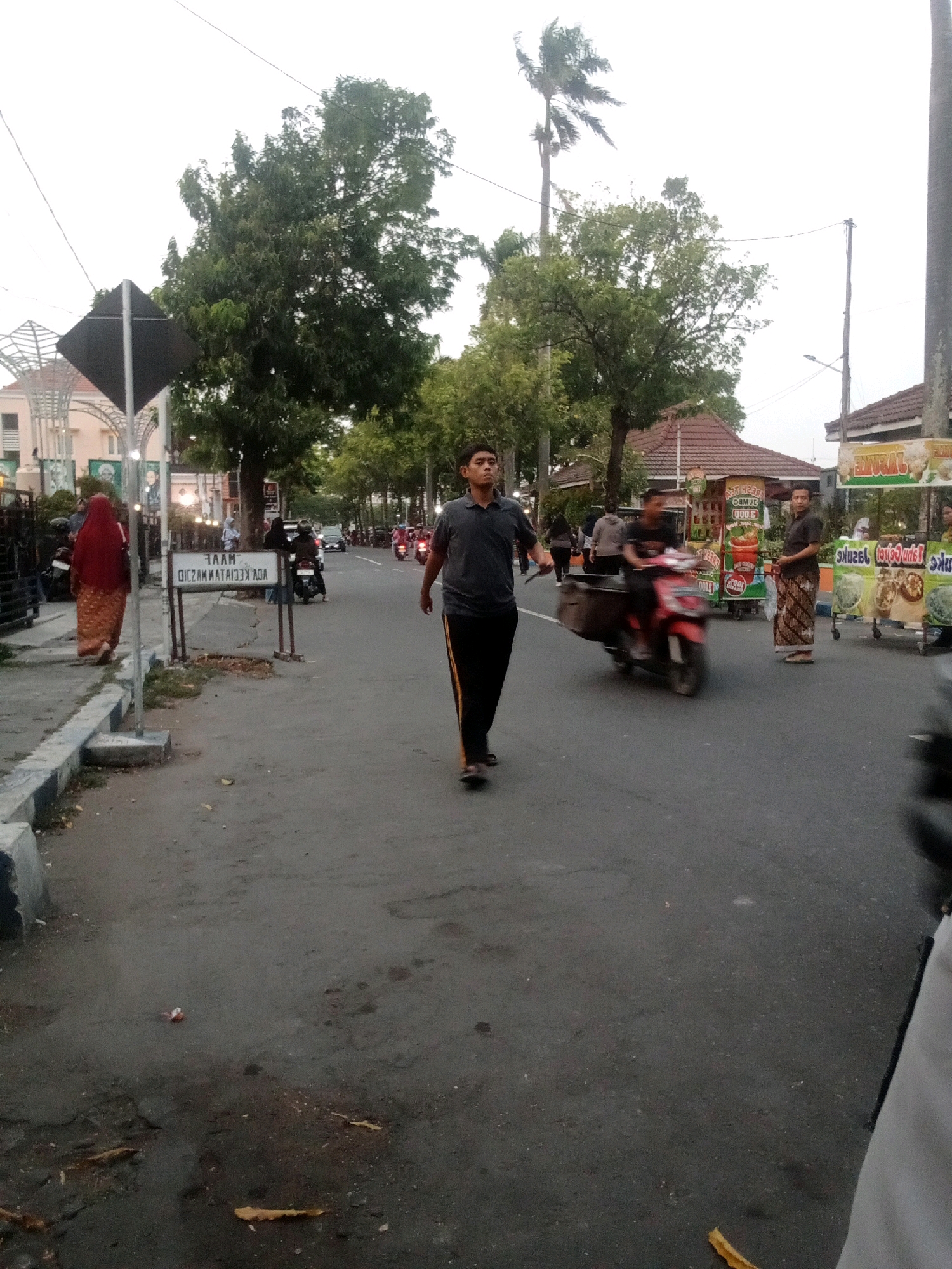 The image depicts a street scene in Jombang, Indonesia, likely during daylight. In the foreground, a young man in a dark gray t-shirt and black pants with a yellow stripe is walking on the asphalt road. He appears to be looking forward. Behind him, a red motorcycle is being ridden by another person, blurred due to motion.
To the left of the road, a sidewalk is visible, bordered by a low concrete barrier with blue and white markings. A woman in a red hijab and patterned orange skirt is walking away from the camera on the sidewalk. A signpost with a dark diamond-shaped sign is also present on the sidewalk.
In the background, several trees line the street, with a tall palm tree on the right. Buildings with reddish-brown tiled roofs are visible, including what appear to be food stalls or small shops. One stall has text that is partially visible and appears to be in Indonesian. Several other people and motorcycles are present in the mid-ground and background, some of whom are also blurred. The sky is overcast. The overall impression is of a moderately busy urban street with pedestrian and vehicular traffic.