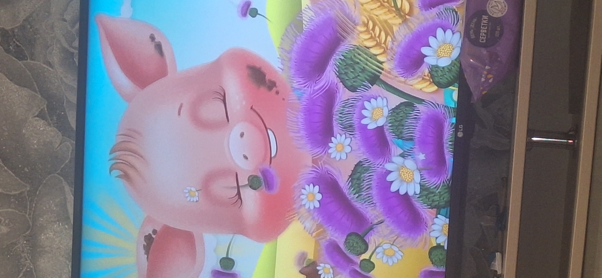 The image depicts a screen displaying a cartoon illustration of a pink pig with its eyes closed and a gentle smile. The pig is adorned with purple thistles and white daisies, with a few dark smudges on its face, suggesting it might be covered in mud. The background features a bright yellow sunburst against a blue sky, with hints of green hills. Below the screen, a dark gray television stand is visible, with a portion of a purple packaging labeled "СЕРВЕТКИ" (which translates to "napkins" in Russian) on the right side. There are no people or animals present in the immediate vicinity of the screen or stand, and the scene does not offer any specific clues that would allow for its location within Bila Tserkva, Ukraine.