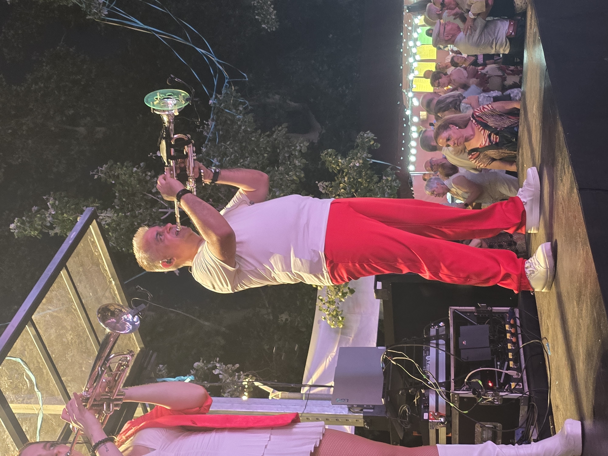 An outdoor nighttime scene in Cornellà de Llobregat, Spain, depicts two musicians performing on an elevated stage for an audience.
On the left, a woman is visible from the mid-torso downwards. She has light skin, dark hair (partially visible), and wears a white short dress with a ruffled hem, a bright red long-sleeved top draped over her shoulders, white fishnet stockings or tights, and white platform boots. She holds a gold-colored trumpet to her mouth, poised as if playing.
To her right, a man is seen from head to toe. He has light skin, short grey hair, and wears a white short-sleeved t-shirt, bright red pants, and white sneakers. An earpiece is visible in his right ear. He holds a silver-colored trumpet, which features a translucent green bell attachment/mute and an attached microphone, and his cheeks are puffed, indicating active playing.
The musicians stand on a dark, elevated stage. To the left side of the stage, an array of professional audio equipment is visible, including stacked racks, a silver laptop or console, a black monitor or speaker, and numerous intertwined cables on the stage floor. Above the stage, a transparent corrugated roof structure supported by metal beams is present.
The background is dark, dominated by dense foliage and trees, indicative of a park or outdoor setting. In the mid-ground, blue ribbons or streamers hang from trees to the right of the stage. In the far background, numerous string lights with small bulbs illuminate a crowd of people, suggesting a festive gathering. Some audience members are facing the stage, while others are conversing. Faint outlines of structures, possibly market stalls or tents with yellow banners, are visible further back. The scene is brightly lit on stage by artificial sources, while the background remains dim.