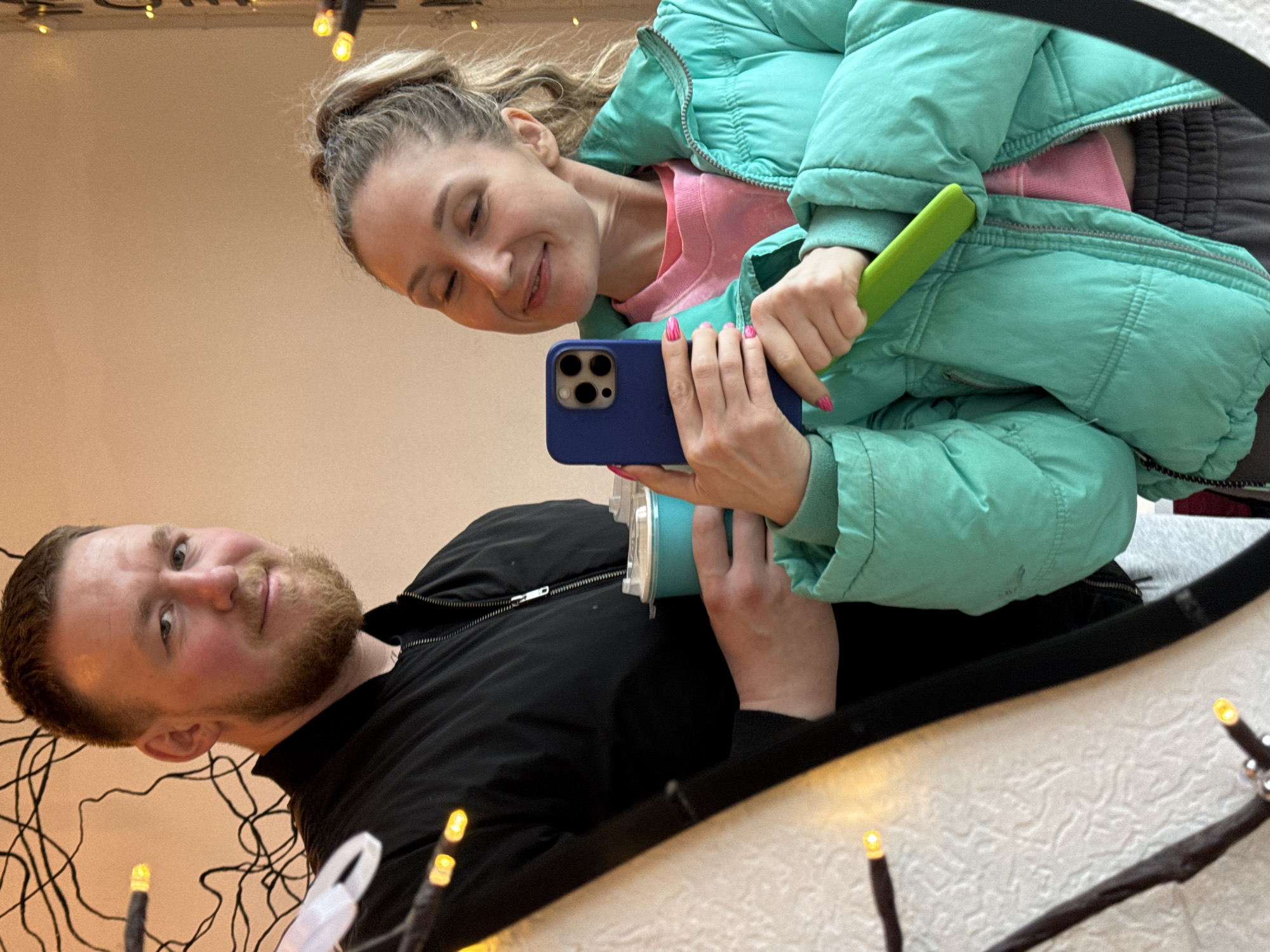 A couple is taking a selfie in front of a mirror. The woman is wearing a light green puffer jacket over a pink top and is holding a blue smartphone. She has her hair in a high bun. The man is wearing a black jacket and has a reddish-brown beard. The mirror frame is visible, and Christmas lights are around the edges. It's impossible to determine that the image was taken in Osa, Russia.