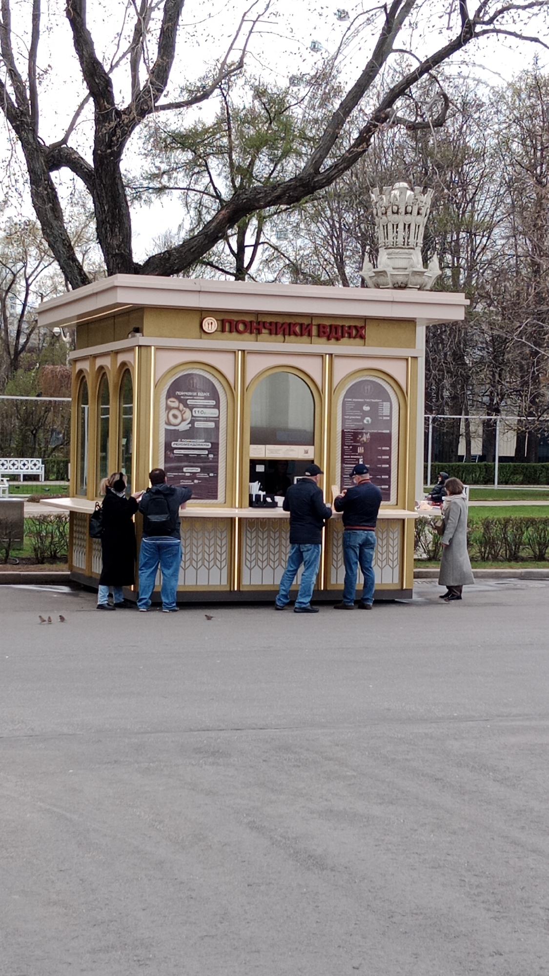 The image shows a donut stand located in VDNKh Park, Moscow, Russia. The stand is made of pink and gold and has the Russian words for "Donuts VDNKh" on the front. There are five people standing in line to purchase donuts. The weather is overcast. There is a tree behind the stand with bare branches. A white park bench can be seen to the left of the stand.