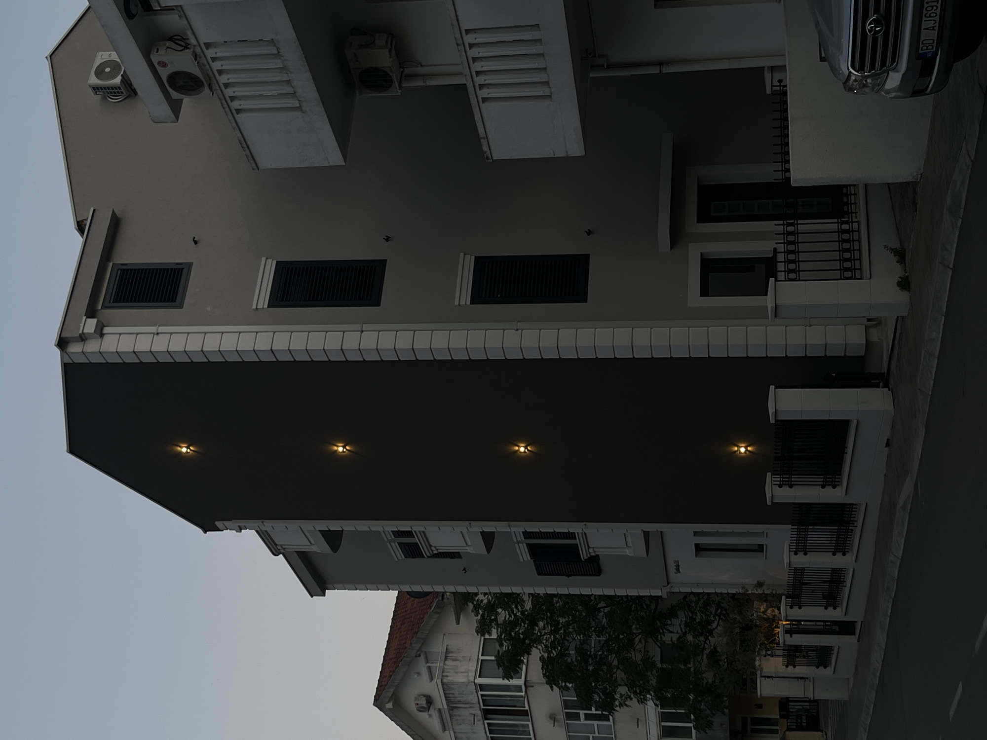The image captures a twilight view of a street in Budva, Montenegro. On the right, a modern, gray building features a dark gray facade with three evenly spaced exterior lights casting a warm glow. The building has dark-shuttered windows and balconies with white railings. To the left of this building is another, lighter-colored building with white decorative trim and dark shutters. Further to the left, a traditional building with a red tiled roof and balconies is visible, with a tree providing a touch of greenery between the buildings. A black metal fence with white-capped pillars runs along the front of the modern gray building, bordering the sidewalk and street. A silver SUV with a license plate reading "BD AJ691" is parked on the right side of the street, partially visible in the foreground. The sky is a pale, clear blue, indicating dusk.