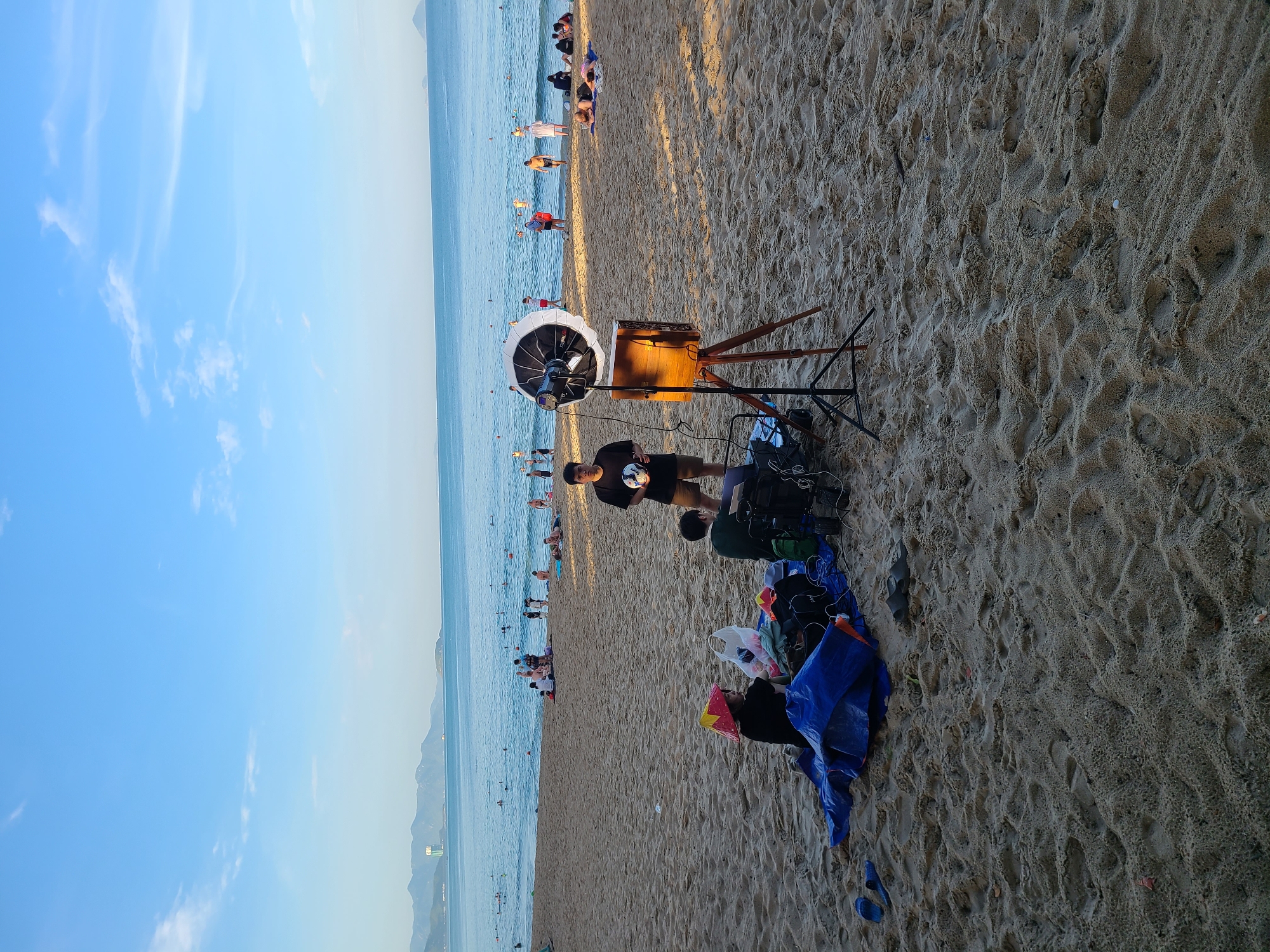 A wide, sandy beach in Nha Trang, Vietnam is bustling with activity. In the foreground, a portable lighting setup with a large softbox on a tripod is positioned next to a wooden box, likely for photography. A young man stands near the equipment, holding a ball. To his left, two individuals are seated on a blue tarp, surrounded by various bags and belongings. In the background, the turquoise ocean stretches to the horizon, dotted with distant hills. Numerous people are scattered across the beach and in the water, some swimming, others relaxing on towels. The sky is a clear, light blue with a few wispy clouds.