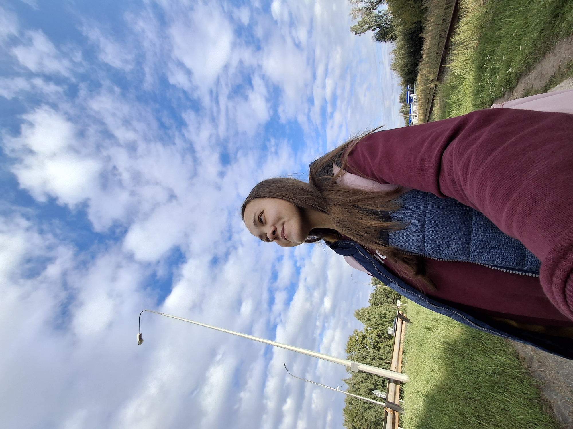 A young woman with long brown hair is standing outdoors, looking up at a partly cloudy blue sky. She is wearing a burgundy sweatshirt and a blue denim vest over it, with a pink backpack visible over her left shoulder. Her right arm is extended towards the camera, suggesting she is taking a selfie. In the background, there are two tall streetlights, trees, a grassy area, a dirt path, and railroad tracks. A building with blue accents and a white roof is visible in the distance beyond the trees and tracks. The overall environment suggests an outdoor, possibly semi-rural or park-like setting. The image appears to have been taken in Chaikovsky, Russia.