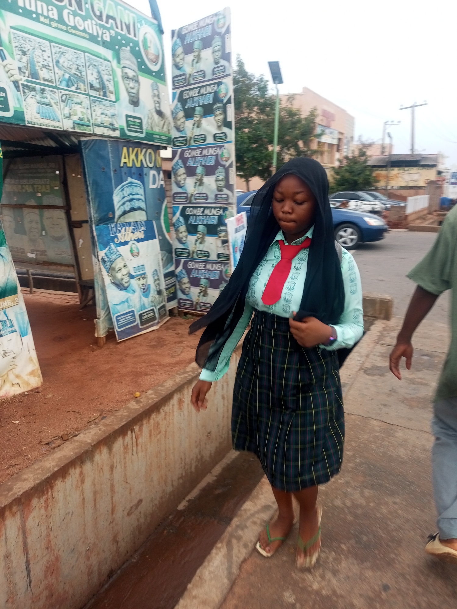 Here's a detailed description of the image:
**People:** A young woman wearing what appears to be a school uniform is walking on a sidewalk. The uniform consists of a light blue button-down shirt, a plaid skirt, a red tie, and a black headscarf. She is wearing flip-flops. The arm and part of the torso of another person are visible on the right side of the frame.
**Location:** The setting is likely a street in Gombe, Nigeria. Political campaign posters are prominently displayed on a structure next to the sidewalk, featuring images of candidates with names such as "Gombe Munga Alkhari" and "Tahan Alheri." These posters provide a contextual clue about the location.
**Objects:** Aside from the clothing and posters, other objects in the scene include the sidewalk, a metal structure supporting the campaign posters, a curb separating the sidewalk from a dirt area, and cars parked along the street in the background.
**Actions/Interactions:** The primary action is the young woman walking along the sidewalk. She is holding the edge of her headscarf. The other person visible is also presumably walking.
**Environment:** The scene appears to be outdoors during the day. The lighting suggests it might be an overcast day.