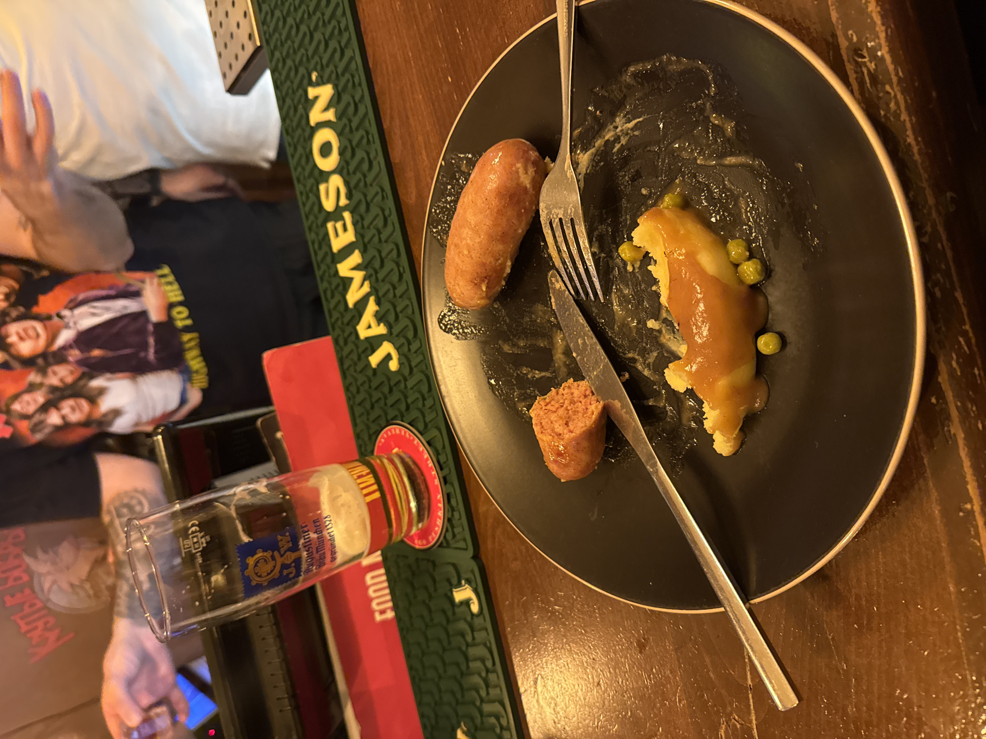 The image shows a partially eaten meal on a black plate. The meal consists of two sausages, mashed potatoes, gravy, and peas. A knife and fork are placed on the plate. Beside the plate is an empty beer glass and a Jameson bar mat. A person is visible on the left of the image, wearing a t-shirt. The setting appears to be a bar or restaurant. The location is Yerevan, Armenia.