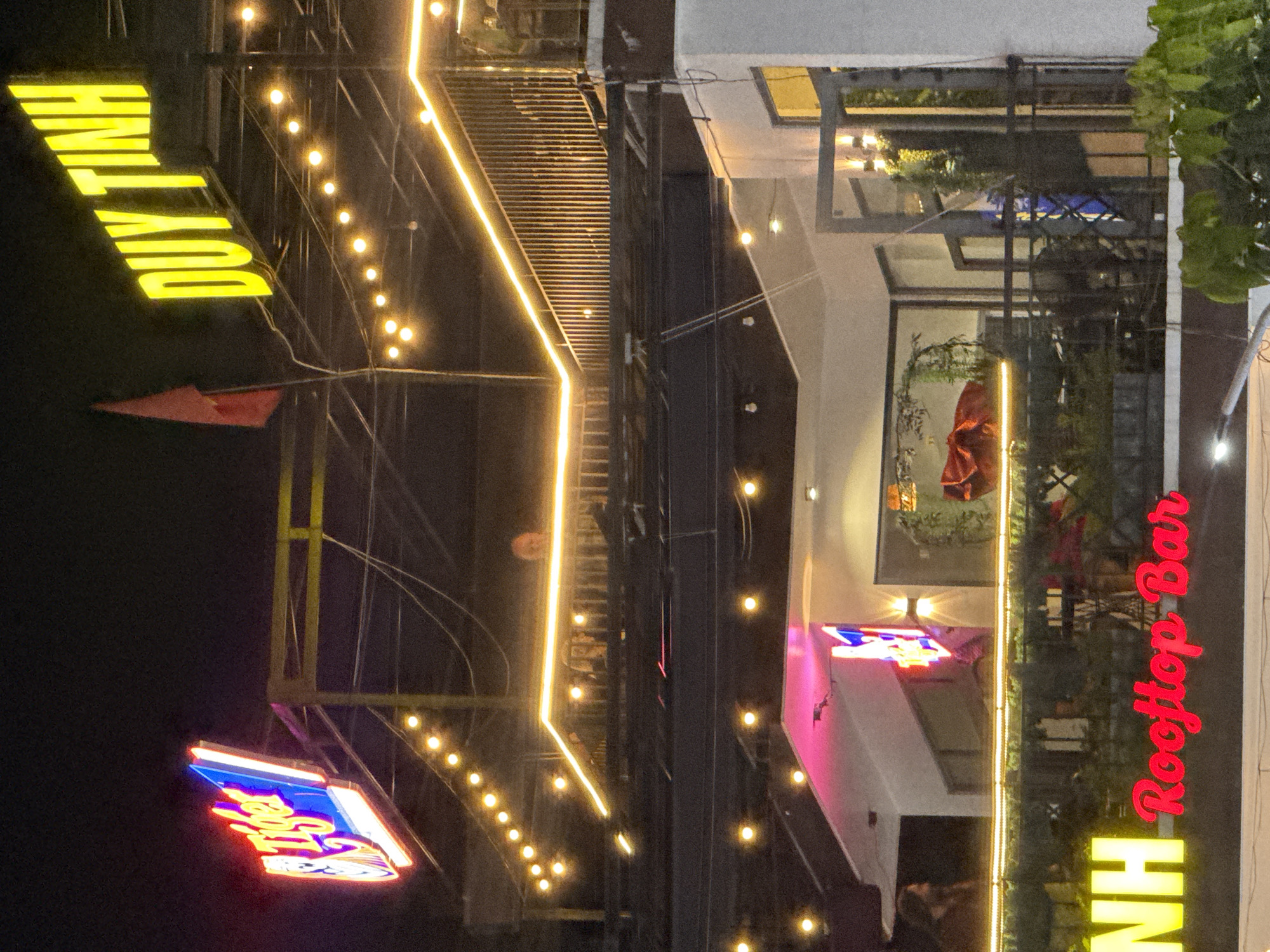 The image depicts a nighttime scene of a rooftop bar in Đà Nẵng, Vietnam. The bar, identified as "NH Rooftop Bar" by its neon red sign, is illuminated by a series of lights. A sign displaying "DUY TINH" in yellow is visible. Another sign, obscured by its angle, features a red and blue neon design. The building appears to be several stories high, with a balcony area visible. The environment is dark, suggesting it is nighttime.
