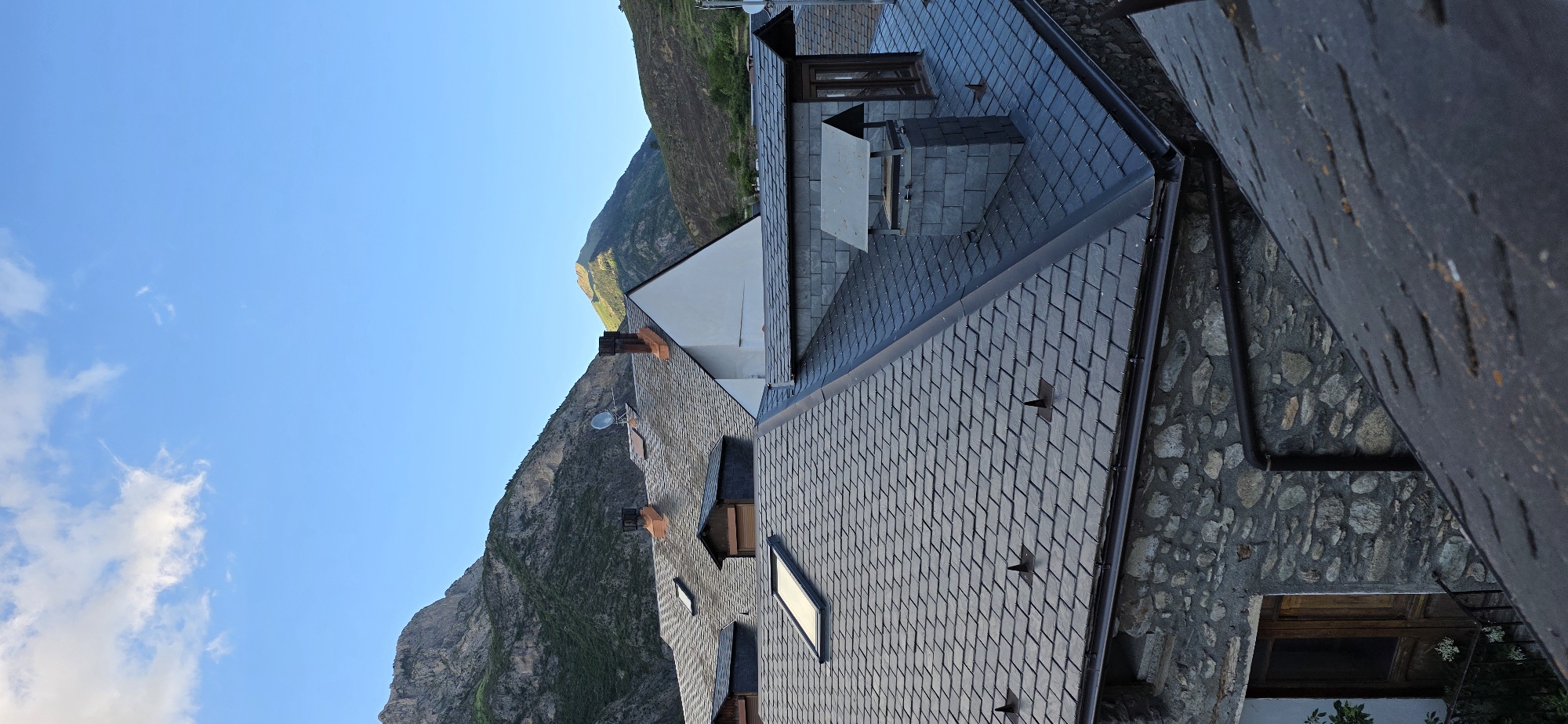 The image captures a scenic view from above the rooftops of traditional buildings, looking towards a majestic mountain range under a clear sky. In the foreground, several buildings are prominent, characterized by dark, interlocking slate tile roofs. These roofs feature brown rain gutters, multiple roof windows or skylights, and terracotta-colored chimneys. One roof also hosts a satellite dish and a larger structure that appears to be an outdoor cooking or ventilation unit. The walls of these buildings are constructed from rugged, irregularly shaped stones, giving them a rustic appearance.
The middle ground is dominated by a grand mountainous landscape, with steep, rocky peaks and slopes interspersed with green vegetation. The lighting on the mountains suggests either late afternoon or early morning, with parts of the peaks illuminated by sunlight and others cast in shadow. The upper portion of the image is filled with a vibrant blue sky, dotted with a few wispy white clouds, indicating clear and pleasant weather.
No people or explicit activities are depicted in the scene. The architecture and natural setting are consistent with a mountainous village environment, such as Benasque, Spain. There is no visible text in the image. The overall impression is one of tranquility and traditional mountain living.