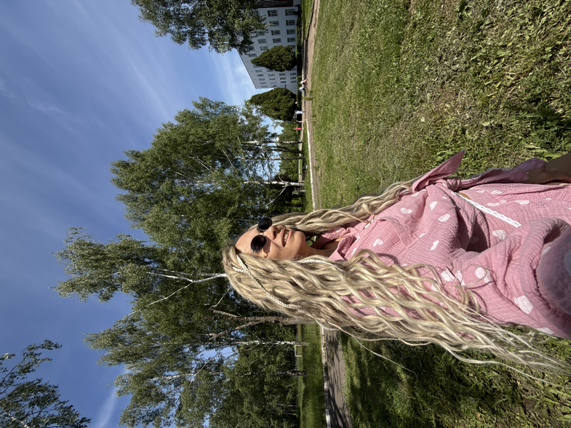 The image displays an outdoor scene on a clear, sunny day in an institutional or park-like setting. In the foreground, an adult woman with long, wavy blonde hair, styled with visible dreadlocks or braids, wears dark, round sunglasses and a pink, textured collared shirt adorned with white heart-shaped patterns. The shirt is tied at her waist, revealing a white lace trim. She is smiling and looking towards the right side of the frame (viewer's left), captured from a low angle, consistent with a selfie.
The mid-ground features an expansive green lawn with patches of varied grass textures and a dark, paved path that curves from the left towards the background. Several tall birch trees with characteristic white bark and green deciduous leaves are prominent, casting shadows on the ground. A faint white goalpost frame is partially visible in the far left-mid ground.
In the background, a large, multi-story white building with numerous rectangular windows stands amidst more trees, including two tall, conical evergreen trees in front of it. Approximately 4-6 small, indistinct figures are visible on or near the paved path in the mid-ground, walking or standing. One figure appears to be wearing red, while others are in lighter attire; one individual seems to be accompanying a stroller or small child. The sky above is a bright, clear blue with faint, wispy white clouds.