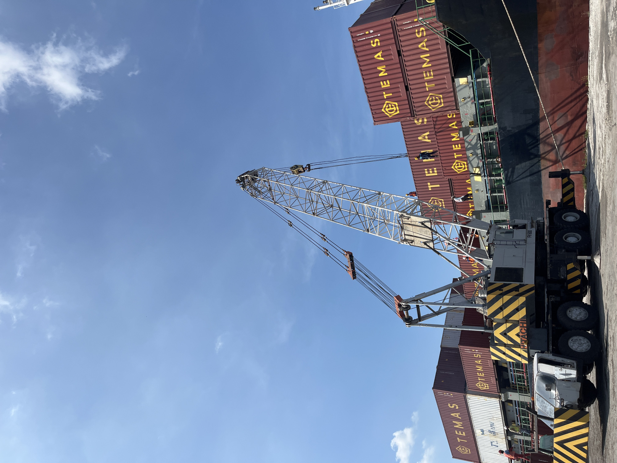 The image depicts a port operation in Tambonbaroh, Indonesia, where cargo containers labeled "TEMAS" are being loaded onto a ship. A large crane, identifiable by its lattice boom and yellow-striped counterweight, is positioned to lift containers. Several workers are visible on top of containers and along the side of the ship. The environment consists of a clear blue sky with scattered clouds. A portion of the ship's hull is visible, showing a red-brown color with signs of wear. The ground is a concrete surface typical of a port loading area.