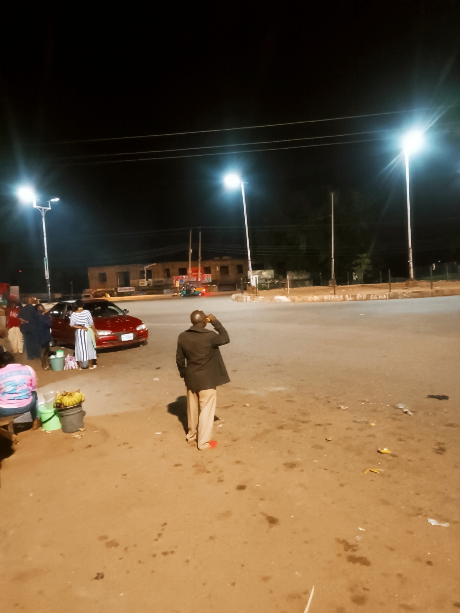 The image shows an outdoor scene at night in Bukuru, Nigeria. Several streetlights illuminate the area. There is a roundabout with a building visible in the background. A red car is parked near the left side of the frame, with several individuals gathered around it. A man in a dark jacket and light-colored trousers stands in the foreground, facing away from the camera. A vendor with a bucket of bananas is on the side of the road. The ground is dirt.