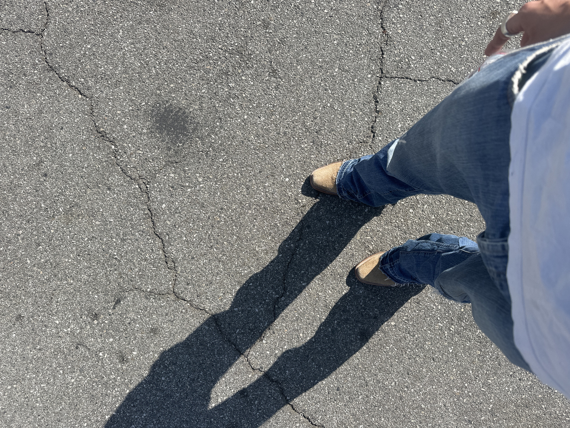 A person stands on asphalt pavement, likely a road or parking lot. The person wears blue jeans and tan suede shoes. A white shirt is partially visible on the right side of the frame. A ring is visible on the person's right hand. Strong shadows cast by the person's legs and feet indicate bright sunlight. Cracks are visible in the asphalt. Based on image analysis, this picture most probably was taken in Glendale, United States of America.