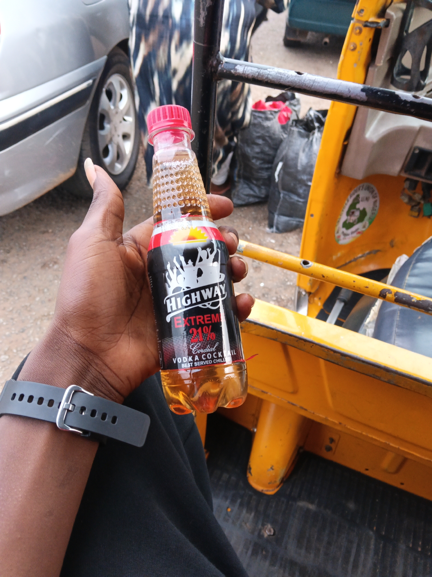 A close-up shot shows a hand holding a bottle of "Highway Extreme" vodka cocktail. The bottle is partially filled with a yellowish liquid and has a red cap. The label on the bottle is black with white lettering and graphics, including the brand name and "21%" indicating alcohol content. A black watch is worn on the wrist. In the background, a yellow auto-rickshaw is visible, along with parts of a silver car and several black bags. The scene appears to be outdoors, possibly in a city or roadside environment.