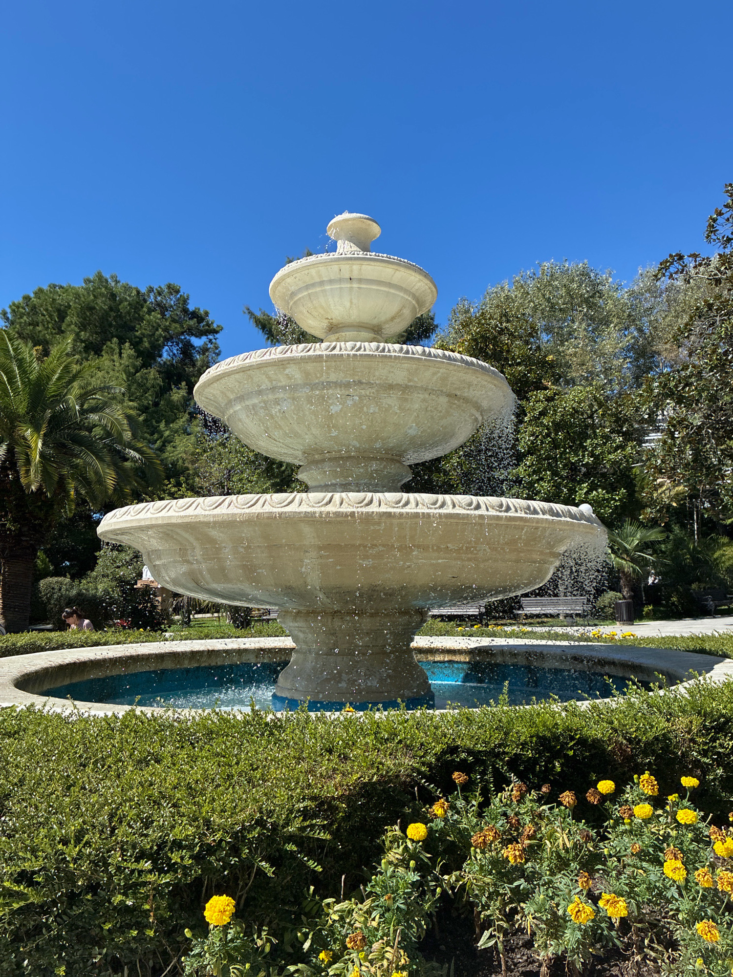 A three-tiered stone fountain stands in the center of a park on a clear day. The fountain is made of light-colored stone and water cascades from each tier into a blue pool at the base. In front of the fountain, a manicured hedge surrounds a bed of yellow flowers. Behind the fountain, trees and palm trees are visible, with a glimpse of benches and a trash bin in the background. A person is seated on the grass to the left of the fountain, near a winding path. The scene appears to be in a park in Sochi, Russia, given the presence of palm trees and the general ambiance of a southern resort town.
