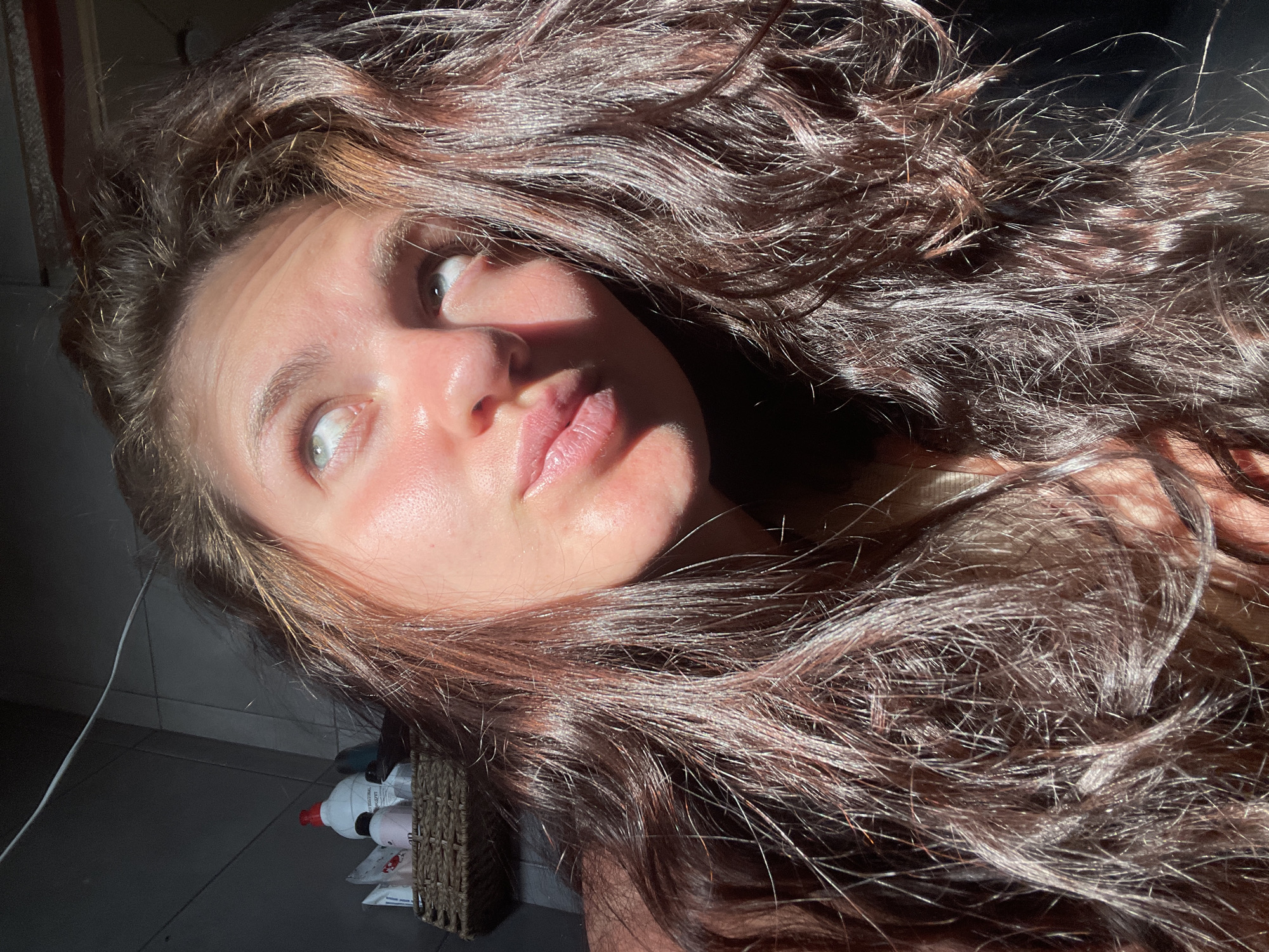 The image shows a close-up, low-angle shot of a young woman's face and hair, bathed in strong sunlight that creates dramatic shadows. She has long, wavy, dark brown hair, with some strands catching the light and appearing reddish. Her skin is illuminated by the sun, highlighting its texture. She is looking upwards and to her right, with her mouth slightly parted. Her eyes are open and appear to be a light blue or grey color, with one eye looking directly into the light.
In the background, to the left of the frame, a woven basket sits on a shelf. The basket contains several bottles, including a white bottle with a red cap and a smaller pink bottle. The wall behind the basket appears to be tiled with dark rectangular tiles. A thin, dark electrical wire is visible running diagonally across the upper portion of the frame. The overall environment appears to be an indoor space, possibly a bathroom or a room with limited natural light sources, given the presence of toiletries and the dramatic interplay of light and shadow. The image does not provide any specific visual cues to pinpoint its location within Мосметаллоконструкция Онт, Russia.