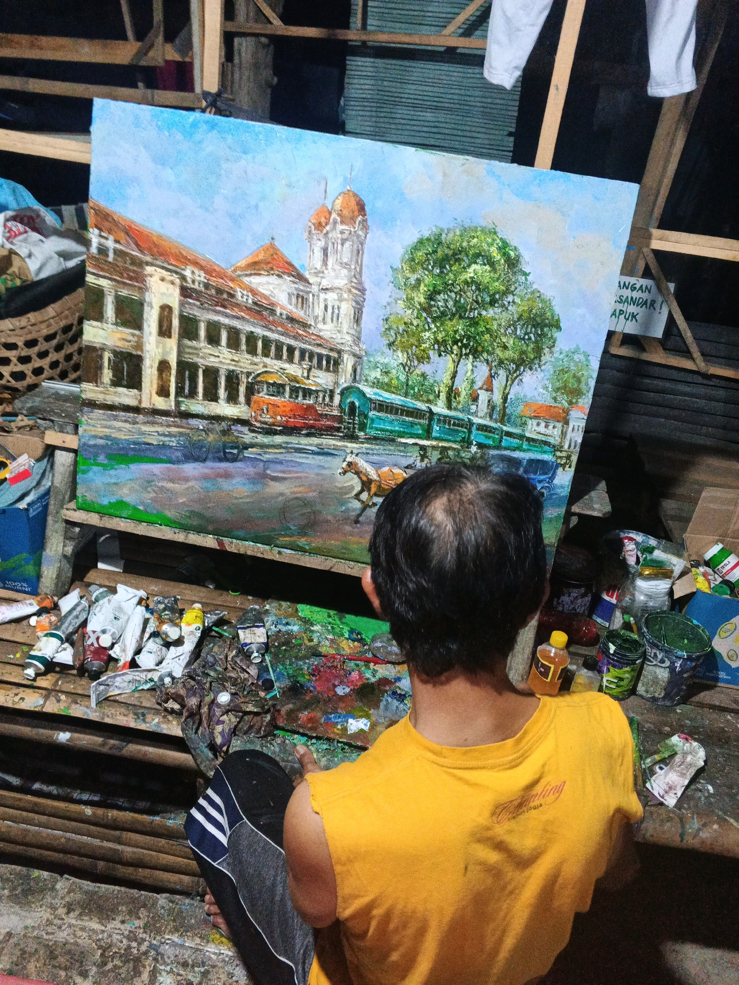 A person wearing a yellow t-shirt and dark pants with white stripes sits on a wooden platform, facing away from the camera and towards a large painting on an easel. The painting depicts a street scene with a colonial-era building, a red tram, a green tram, and a horse pulling a carriage. Trees and a blue sky with clouds are also visible in the painting. The platform is cluttered with art supplies, including tubes of paint, brushes, and containers of paint. A woven basket filled with what appears to be fabric is situated to the left of the painting. To the right of the painting, a sign is attached to the easel, reading "JANGAN SANDAR! LAPUK". The background of the image consists of wooden beams and what looks like a corrugated metal wall, suggesting an outdoor or workshop setting. The overall impression is that of an artist at work in a rustic environment, possibly in Ungaran, Indonesia, given the contextual information.