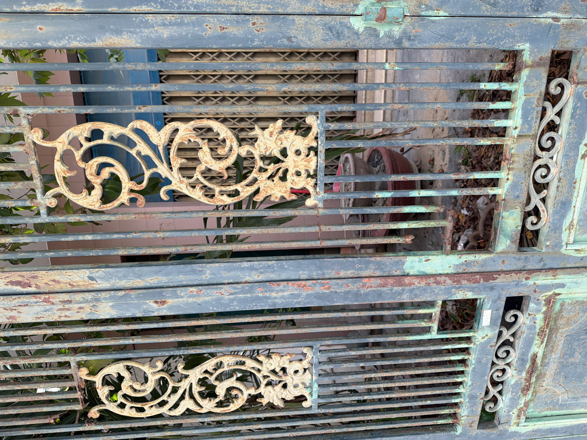 The image shows a rusted, weathered metal gate in front of a building. The gate has ornate decorative elements that appear to be made of wrought iron and are painted a light color, possibly white or cream, with rust showing through. The main structure of the gate is made of vertical metal bars painted in a muted blue or teal color, with significant areas of rust and peeling paint visible. Behind the gate, a darker colored metal collapsible gate can be seen, suggesting a secure entrance. To the left of the main gate, there is a partial view of a building with pink walls and a blue collapsible gate. Some foliage and large potted plants are visible behind the main gate, and the ground appears to be covered in debris, including leaves and plastic. The overall impression is one of age and neglect. The setting is likely an outdoor urban environment.