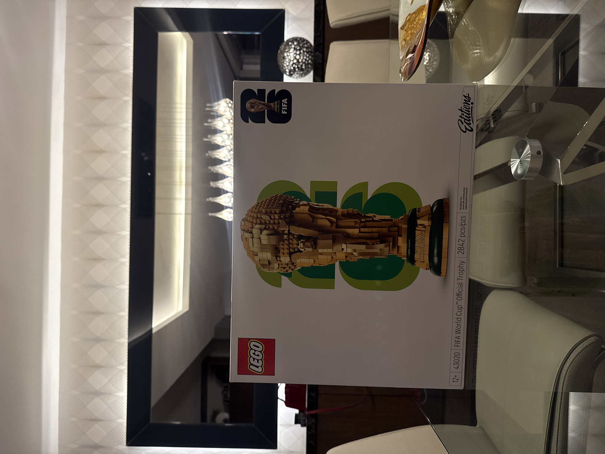 Here is a detailed description of the image:
**Scene:** The image shows a LEGO set of the FIFA World Cup trophy placed on a glass table. A part of a white chair and a decorative sphere are visible. Behind the table is a black-framed mirror or artwork with internal lighting, adding depth to the scene. The left side of the frame reveals a textured wallpaper or wall covering.
**Objects:**
* LEGO set box of the FIFA World Cup trophy.
* Glass table.
* White chair.
* Decorative sphere.
* Black-framed mirror or artwork with internal lighting.
**Text Elements:**
* Visible text on the LEGO box includes the LEGO logo, FIFA logo, "FIFA World Cup™ Official Trophy", "2842 pcs/pzs", "Building Toy", and "Juguete para Construir".
* "Editions" is printed on the right side of the box.
**Environment:** The setting is indoors, likely in a modern or contemporary living space given the furniture and decorations.