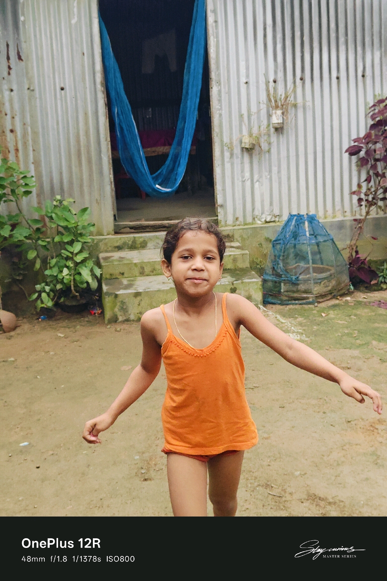 A young girl with curly hair, wearing an orange tank top and red shorts, stands in a yard. She has a thin gold chain around her neck and small gold hoop earrings. She is looking directly at the camera with a slight smile. Her left arm is bent at the elbow, and her right arm is extended to her right. Behind her, a corrugated metal structure with an opening reveals a blue hammock hanging inside. Steps lead up to the opening. To the right of the opening, a blue net covers a round object. The yard appears to be dirt with patches of grass. The scene is likely outdoors in a rural setting. The lighting suggests it is daytime, possibly overcast or late afternoon. The text at the bottom of the image indicates the device used for the photograph: "OnePlus 12R 48mm f/1.8 1/1378s ISO800" and a signature that reads "Stay curious" with "MASTER SERIES" below it. The location is identified as Pipulbari Pt.IV, India.