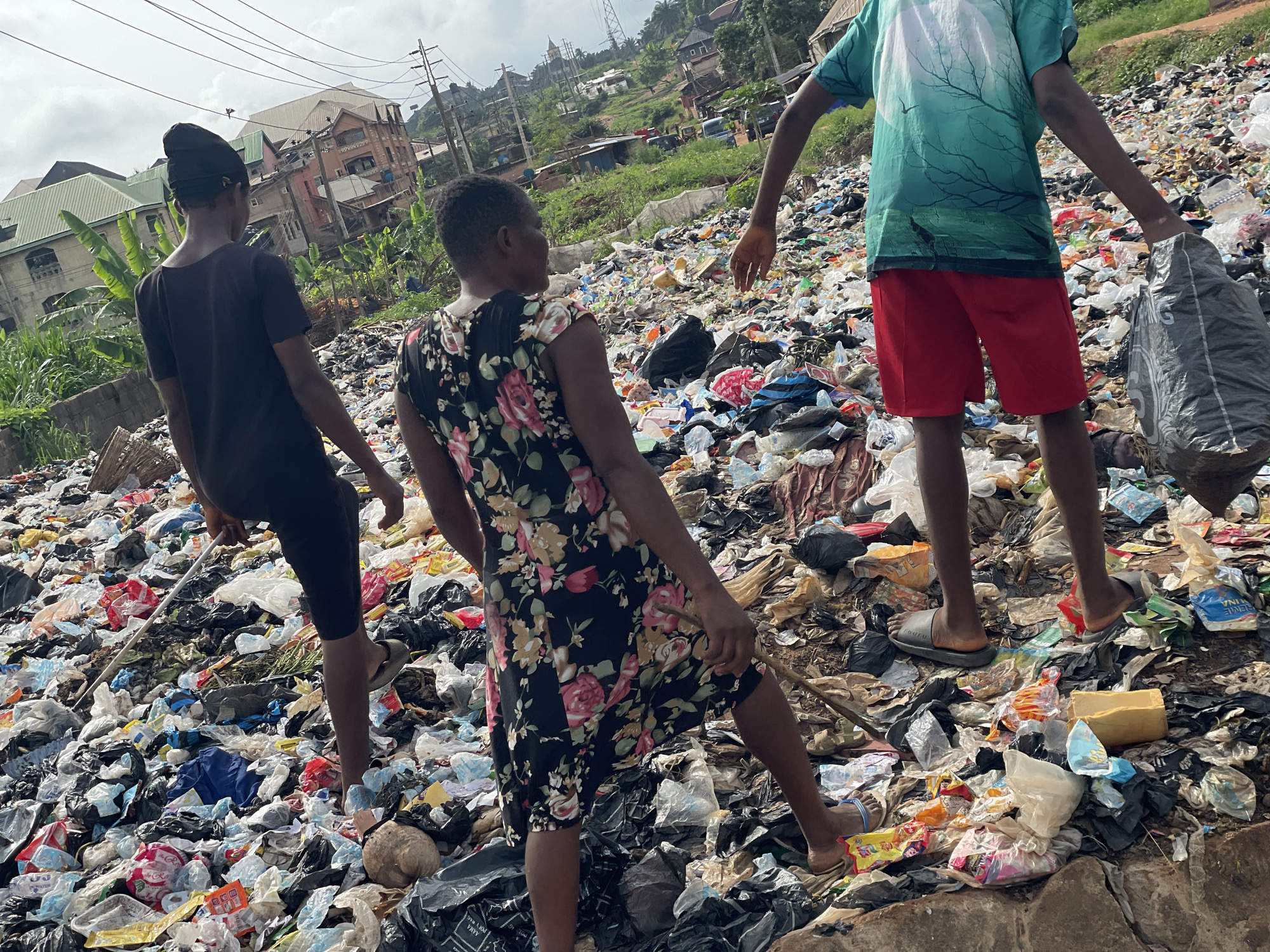 Three people are seen walking in a large trash heap in Nkpor, Nigeria. The trash consists of plastic bottles, bags, food containers, and other discarded materials. The person on the left wears a dark shirt and pants, while the person in the middle wears a dark floral dress. The person on the right wears a green shirt, red shorts, and carries a large bag. Houses and buildings can be seen in the background.