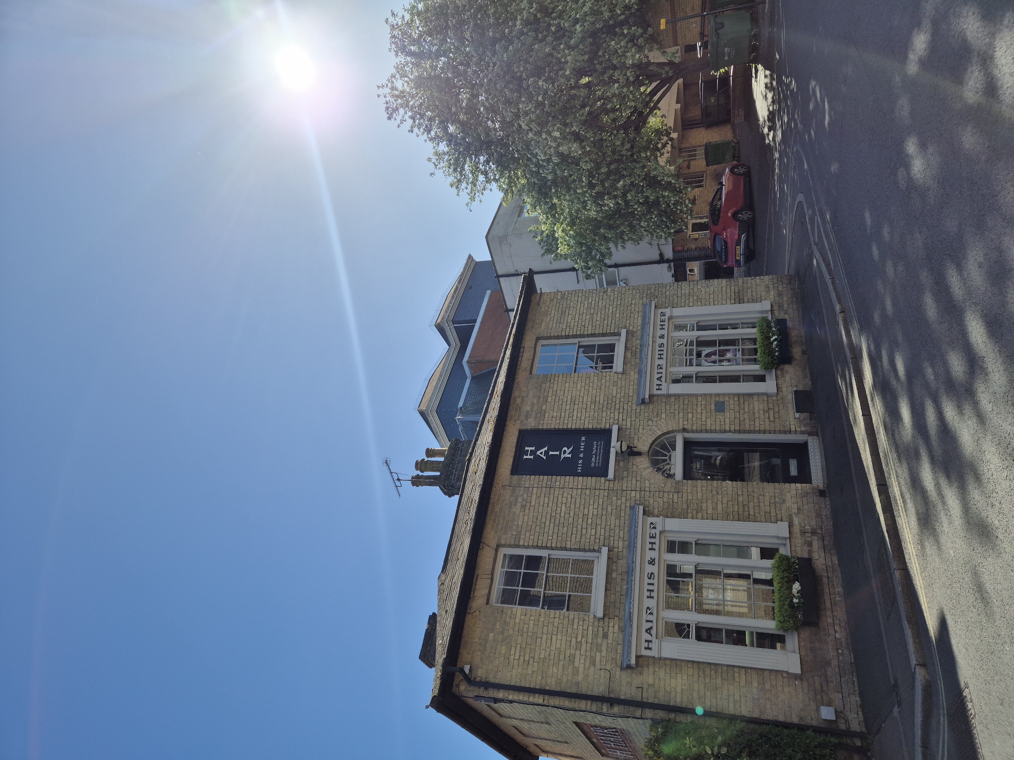 A bright, sunny daytime image captures a street scene in what appears to be West Suffolk, United Kingdom. The primary subject is a two-story building constructed of light-colored brick, situated alongside a paved road. The building houses a hair salon named "HAIR HIS & HER," prominently displayed on several signs.
Above the ground-floor windows, "HAIR HIS & HER" is spelled out in white letters on a dark background. Another black sign, positioned higher on the building, also reads "H A I R" vertically with "HIS & HER" horizontally below, alongside a phone number ("01284 750909") and a website ("www.hairhisandher.co.uk"). The building features multiple white-framed windows; some on the ground floor have decorative window boxes filled with green foliage. A dark main entrance door is visible below a round-arched window. The roof is dark, and an antenna is visible on the chimney.
To the left of the building, a large, leafy tree casts dappled shadows across the road. A red car is parked on the side of the road further down. The sky is a clear, bright blue, dominated by a brilliant sun emitting strong lens flares in the upper left quadrant of the image. The sunlight suggests a clear, warm day. No people are visible in the scene. The architecture and road markings are consistent with a British setting.
