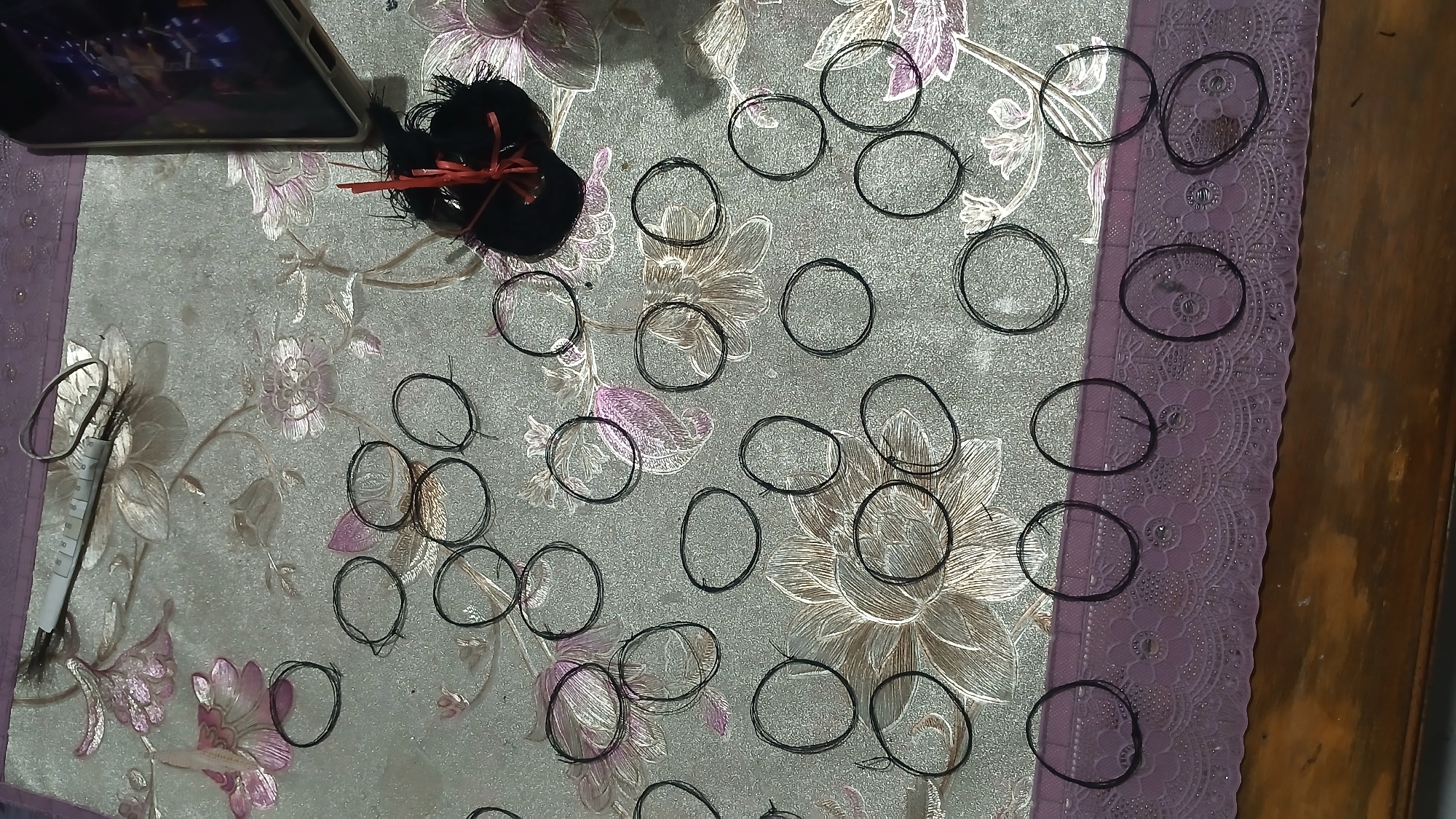 The image shows a close-up, top-down view of a patterned tablecloth with multiple small black elastic bands scattered across it. The tablecloth has a floral design in shades of pink and white against a light gray background. In the upper left corner, a white object with a measuring tape wrapped around it lies next to a coiled wire. To the right of this, a black mass with red threads sticking out is visible, and next to it, a smartphone is partially visible. The elastic bands are mostly circular, with some appearing slightly flattened or distorted. Some bands are isolated, while others are clustered together. A purple lace trim runs along the bottom edge of the tablecloth. The lighting suggests an indoor setting, possibly a table in a home. There is no information in the image that can be used to determine the location in the city of Sampang, Indonesia.