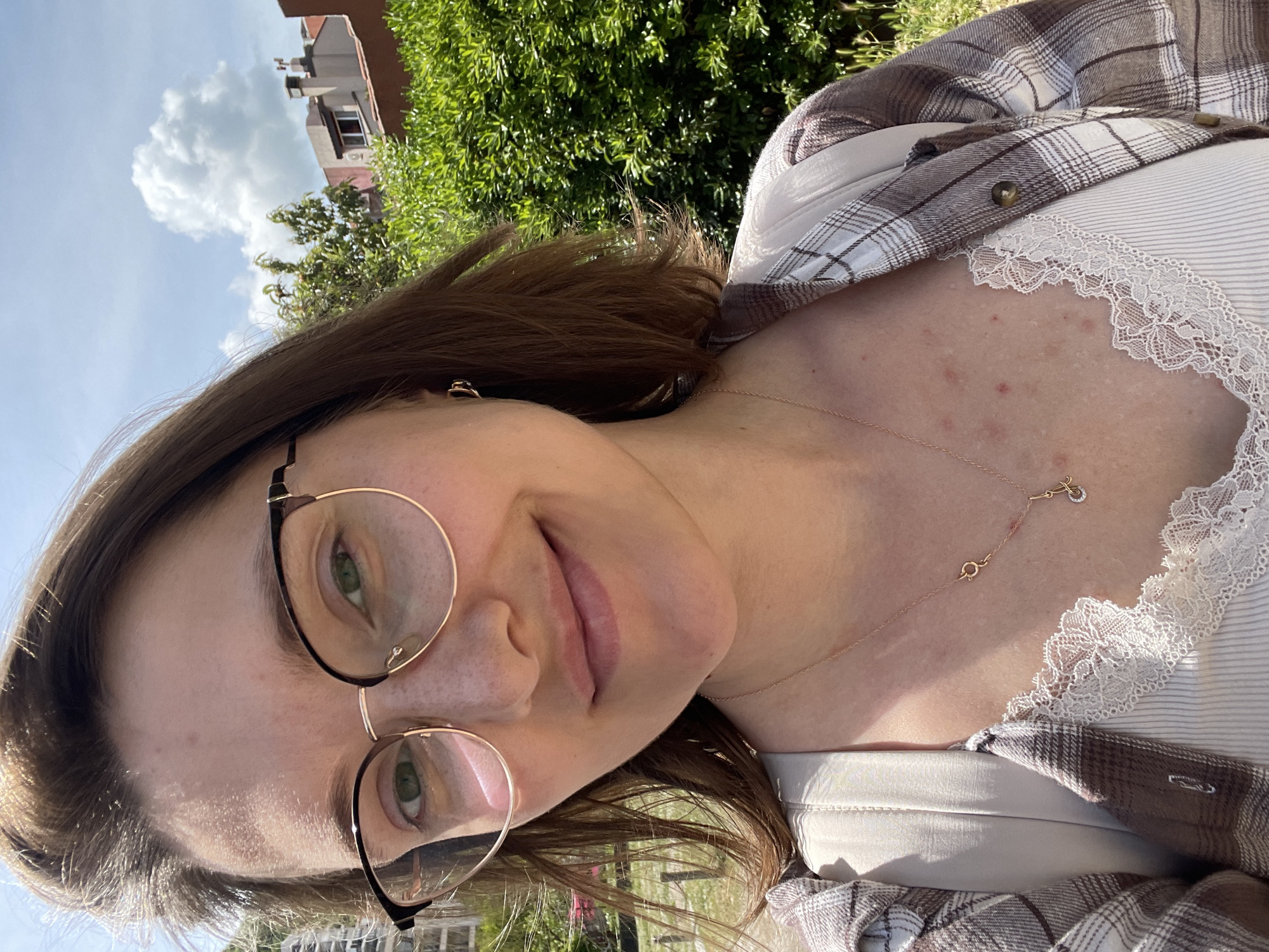 The image is a selfie of a woman, likely taken outdoors in a sunny location. She has light skin, brown hair, and green eyes, and wears round eyeglasses. She is wearing a white tank top with lace trim and a brown and white plaid shirt. A gold necklace is visible around her neck.
The background includes green foliage, suggesting trees or bushes, and a building with a red roof is partially visible. Given the mention of Budva, Montenegro, it is reasonable to assume this image was taken in or near that city, which is known for its coastal landscape and urban architecture. The presence of a building, trees, and clear skies suggests an outdoor location within the city limits or a nearby area.