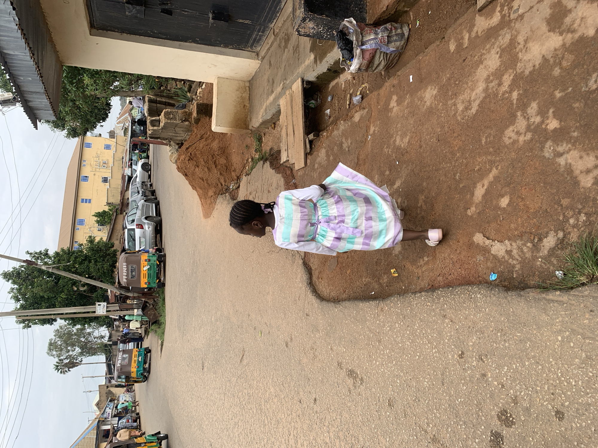 A young Black girl, viewed from the rear, walks on a reddish-brown dirt path adjacent to a paved road in Jos, Nigeria. She wears a long-sleeved white shirt beneath a striped dress featuring horizontal bands of white, light purple, and light aqua/mint green, cinched at the waist with a purple ribbon. Her hair is braided with small purple ties, and she wears light pink sandals.
To her right, a concrete-lined drainage ditch containing litter is partially covered by two wooden planks. A large, open sack filled with black trash bags rests next to the ditch. A light-colored building with a dark wooden door is visible on the far right. A pile of reddish-brown sand is present on the dirt path.
The paved road on the left features several tricycles, common in Nigeria, including one marked "PTA-1723" and another "PT19210" on their rear canopies. Other cars and people are visible along the road. In the background, buildings line the street, notably a multi-story yellow building with blue-framed windows. Utility poles with overhead wires, as well as various trees and palm trees, are scattered throughout the scene under an overcast sky.