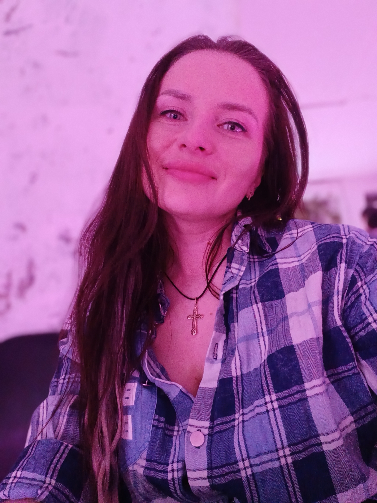 A woman with long brown hair is looking at the camera with a slight smile. She is wearing a blue and white plaid shirt and a necklace with a cross. The lighting in the image has a pink tint. The location is not discernible from the image.