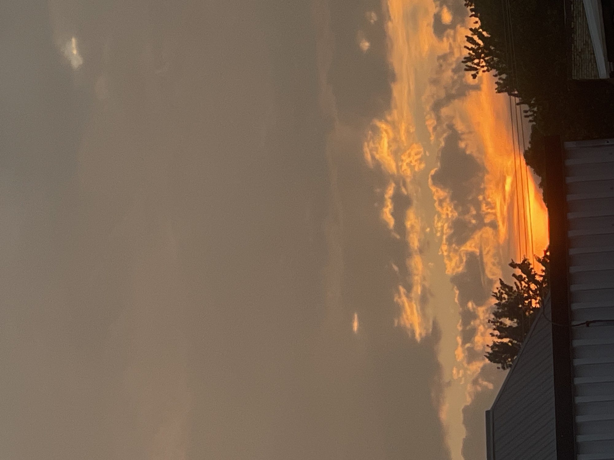 A sunset is depicted over Bluewater, Canada. The sky is filled with a mixture of gray and orange clouds, with the sun's rays creating a vibrant glow. Silhouetted trees and the roof of a building are visible in the foreground. The overall scene is a natural landscape at dusk.