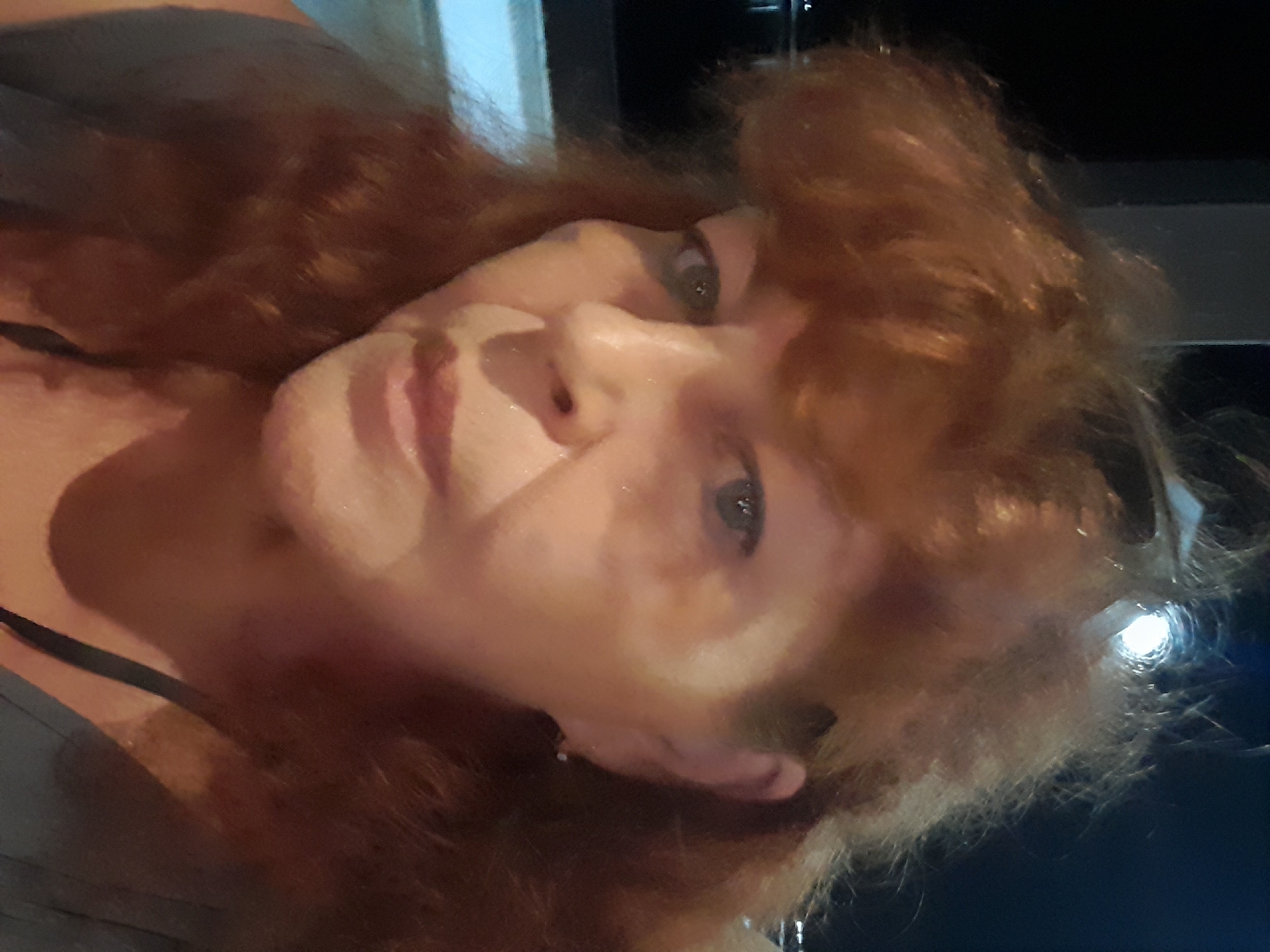 A selfie of a fair-skinned person with reddish-brown hair, angled slightly downward. The background is indistinct, showing dark areas and some reflective surfaces. The person's face is the primary focus, revealing their eyes, nose, and mouth. Their hair is styled with loose curls and appears voluminous. A thin strap, possibly from clothing, is visible on their shoulder. There's no evidence to locate the setting in บ้านบางรักษ์, Thailand.