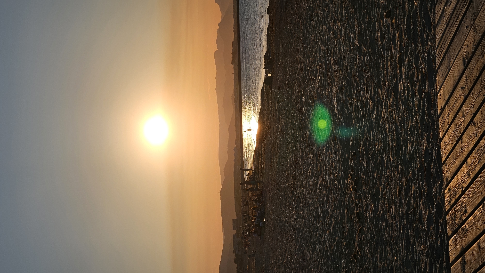 This is a vertical, eye-level, full shot of a beach at sunset in Deveses, Spain. The sun is low in the sky, casting a warm, orange glow over the scene. The sun is a bright, white orb in the upper center of the image. The sky above the horizon is a gradient of light blue to pale orange, transitioning to a deeper orange closer to the sun. The ocean is calm and reflects the sun's light, creating a shimmering path across the water.
In the middle ground, a sandy beach is populated with several groups of people. Some are sitting or lying on the sand, while others are standing or walking. Several individuals are in the shallow water, with a few people swimming. The coastline stretches into the distance, with hazy, layered mountains forming a backdrop. Buildings are visible along the distant shore.
The foreground is dominated by the textured surface of the sandy beach. In the lower right corner, a portion of a wooden boardwalk or pier is visible, with planks running diagonally. A green lens flare is present in the sand in the mid-ground, slightly to the left of the center. The overall atmosphere is tranquil and serene, capturing the beauty of a sunset at the coast.