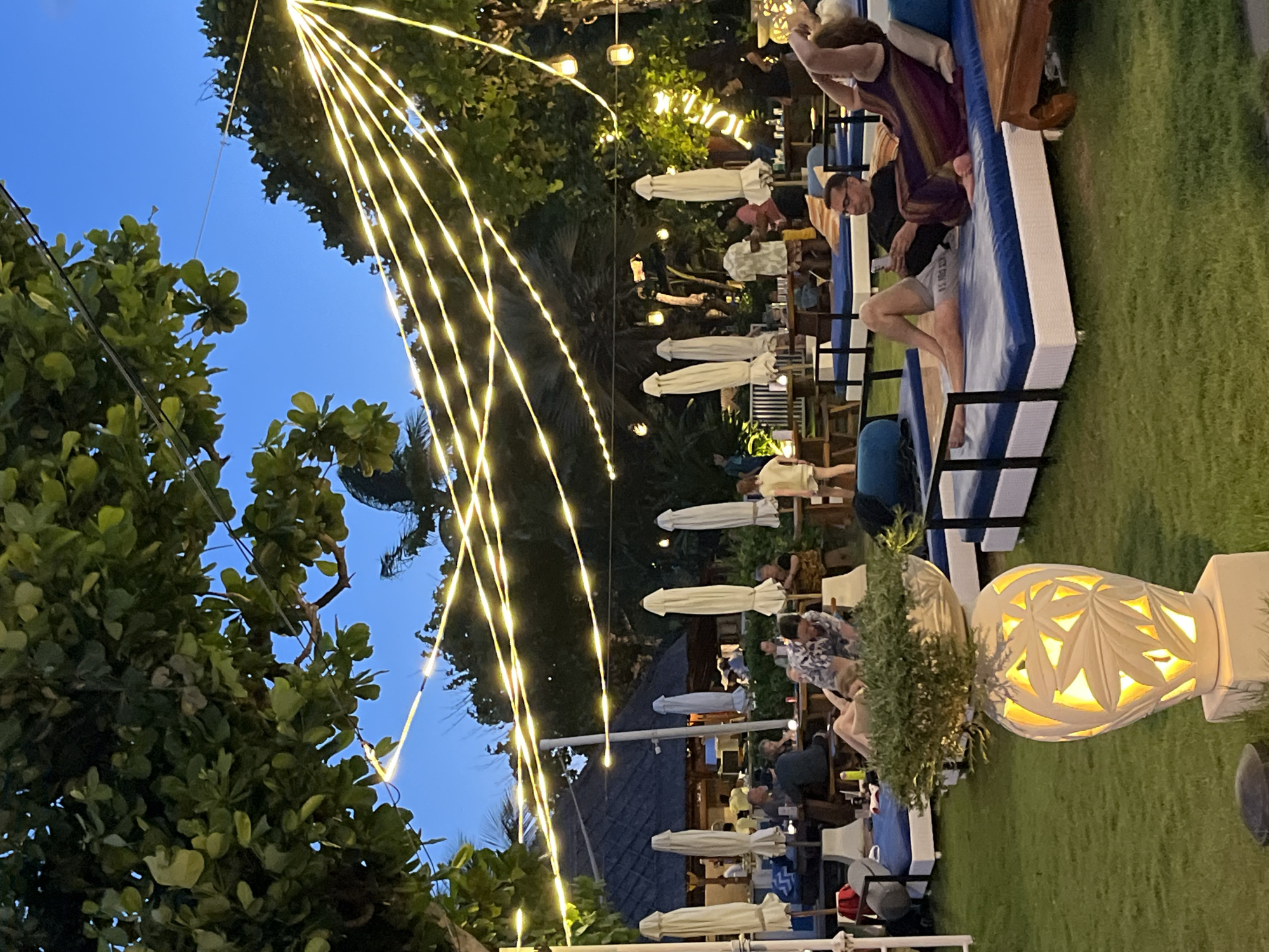 This outdoor scene in an unidentified location in Unknown, Indonesia shows a leisure area with several people enjoying a relaxed atmosphere. The time is likely dusk or early evening, given the lighting. Strings of decorative lights are strung above, curving down from the trees and adding ambiance. The area appears to be a restaurant or lounge, featuring covered tables and chairs. Several individuals are seated at these tables, engaged in what appear to be conversations and possibly dining. The setting includes greenery, with trees and grass visible. Additionally, decorative lighting fixtures are placed along the ground, adding to the overall illumination. A lounge area with padded seating is visible towards the right of the image.