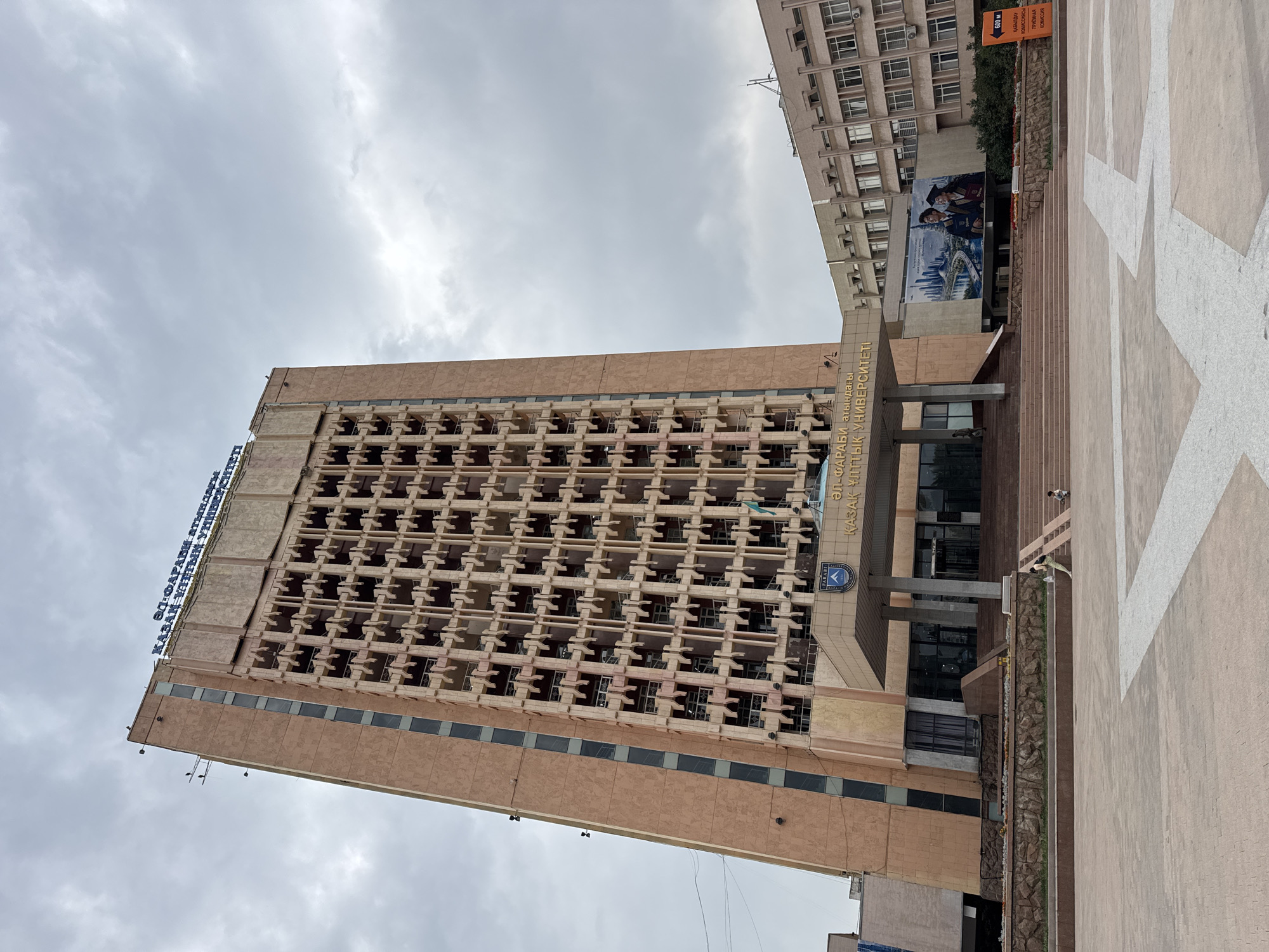 This is a full-frame, eye-level, outdoor, daylight photograph of the Al-Farabi Kazakh National University building in Almaty, Kazakhstan. The dominant structure is a tall, monolithic, Brutalist-style building with a pinkish-brown facade and a repetitive grid of small, recessed windows. The top of the building features blue lettering that reads "ӘЛ-ФАРАБИ атындағы ҚАЗАҚ ҰЛТТЫҚ УНИВЕРСИТЕТІ" (Al-Farabi Kazakh National University). Below this, a cantilevered entrance structure is visible, adorned with a coat of arms and more Kazakh text. The entrance is reached by a wide set of stairs. To the right of the main building, a lower, lighter-colored building with a grid of windows is partially visible. A large mural depicting a modern city skyline and two graduates in academic regalia is mounted on this side building. In the foreground, a paved plaza with a geometric pattern is visible. Two individuals, an adult and a child, are present in the plaza. The sky is overcast with gray clouds.
