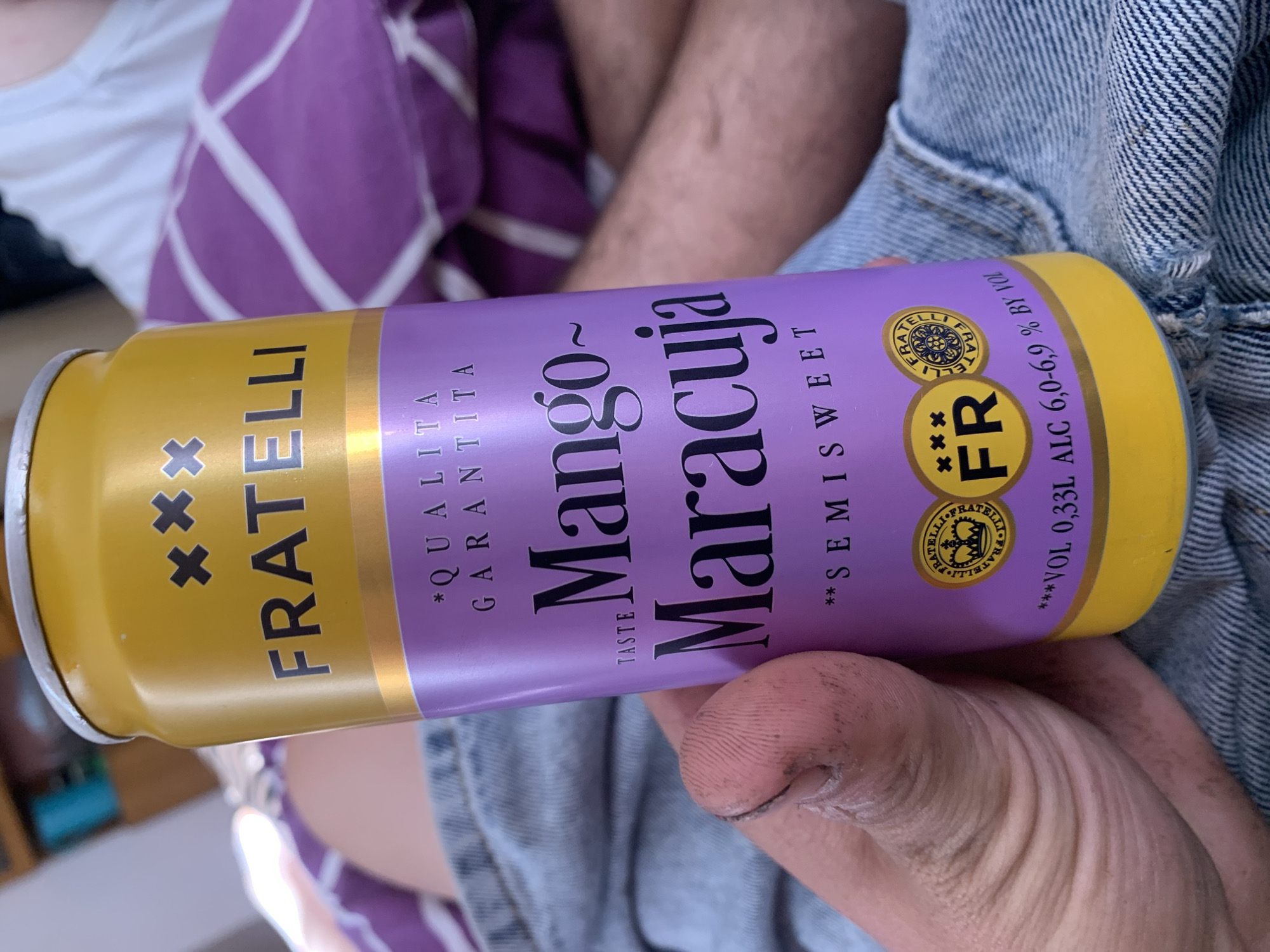 The image shows a close-up of a person's hand holding a yellow and purple can of "FRATELLI Mango-Maracuja" semi-sweet drink. The can has branding that includes the word "FRATELLI" in large black letters and symbols. Below that, it reads "*QUALITA GARANTITA" and "TASTE Mango-Maracuja". Further down, it says "**SEMISWEET" and shows a circular emblem with text and a crown, followed by "FR". The bottom of the can has "**VOL 0,33L ALC 6,0-6,9% BY VOL". The person's hand is visible in the foreground, with dirt on the fingers. The background is blurred but shows a person lying down, wearing a white shirt and a purple and white striped blanket or fabric. The scene appears to be indoors, possibly a home environment. There is no indication of the location being in Zhmerynka, Ukraine.