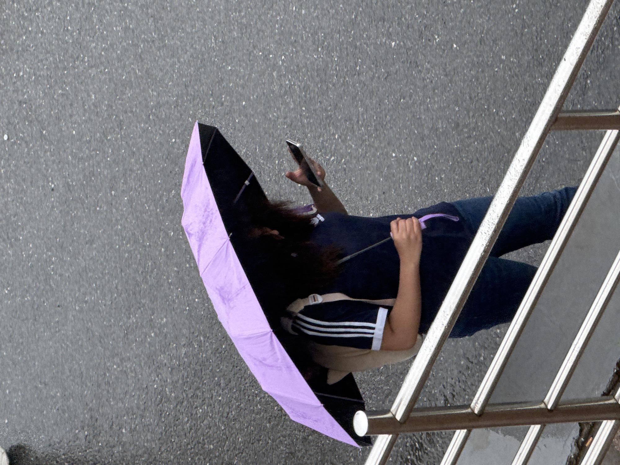 A top-down view depicts a person standing on a wet, grey concrete surface under a partially opened purple and black umbrella. The person is holding a smartphone in their left hand and appears to be looking at the screen. They are wearing dark blue jeans, a dark blue t-shirt with white stripes on the sleeves, and a light-colored backpack with white straps. A metal handrail is visible to the right of the person, suggesting an elevated position. The scene is located in Zhongtun, China.