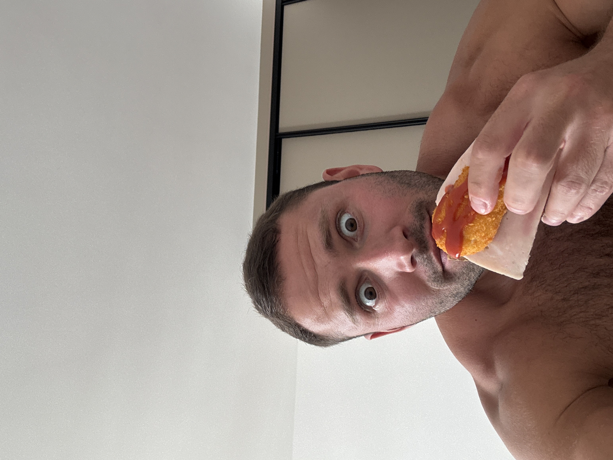 A close-up, low-angle, indoor selfie shows a shirtless man with wide eyes and an open mouth holding a piece of food to his lips. The food appears to be a chicken nugget covered in ketchup, placed on a slice of deli meat. The man has short brown hair and stubble. He is in focus, with the background slightly blurred. The background consists of a white wall and a closet with sliding doors. The lighting suggests an indoor setting, possibly a bedroom, with natural light coming from the left. The man's musculature is visible. The overall impression is that of a spontaneous, perhaps humorous, selfie. There are no visible indicators of the specific location in Budva, Montenegro, or any other city.