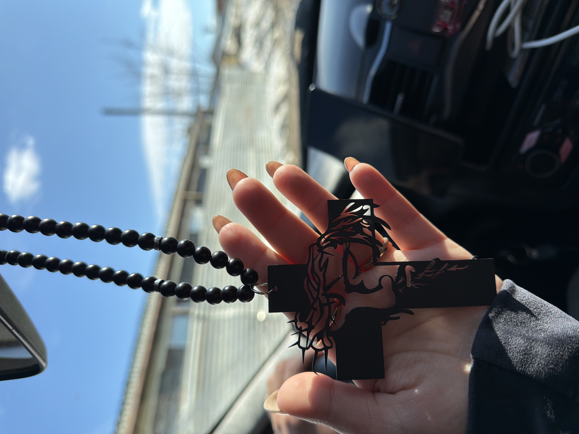 A person is holding a black cross pendant with a black bead necklace attached. The pendant has an intricate design depicting what appears to be Jesus Christ. The person's hand has long, manicured nails. The background is blurred but shows a partly cloudy sky and what seems to be an urban setting with buildings. The photo appears to be taken inside a car. The location is assumed to be the city of Chaikovsky, Russia.