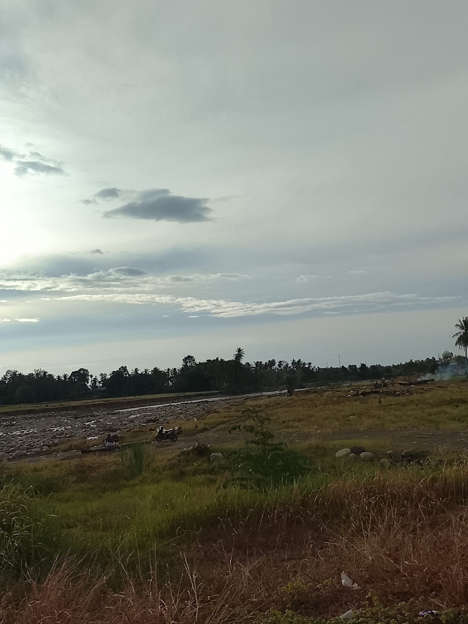 The image depicts a broad, slightly overcast sky with scattered clouds over a rural landscape. The foreground consists of dry, tall grass, suggesting a dry season or arid conditions. Behind the grass is a field with sparse vegetation and a rocky riverbed or dry river channel. In the middle ground, a line of trees forms a dense boundary. Several people are visible near the riverbed, and a motorcycle is parked on a dirt path. Further to the right, a palm tree stands tall, and a small amount of smoke is rising, possibly from a fire. The overall scene appears to be in Geunteng, Indonesia, a location known for its natural scenery and agricultural areas.