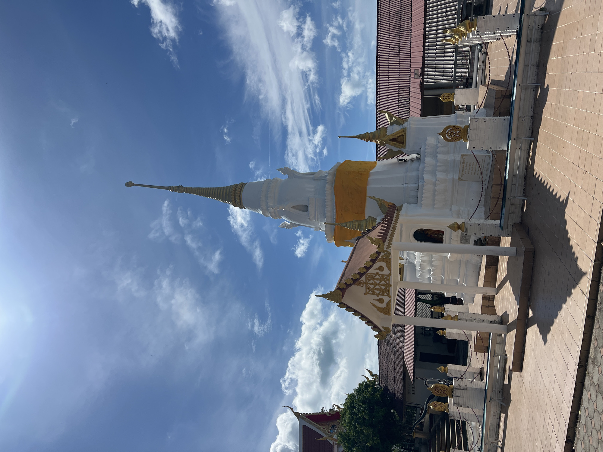 A bright, sunny day illuminates a white stupa with a golden spire at a temple in Baan Hua Thanon, Thailand. The stupa is adorned with a yellow sash and surrounded by a low white fence with golden finials. A small white pavilion with a decorative roof stands in front of the stupa. In the background, red-tiled roofs of other temple buildings are visible, and a lush green tree grows to the left. The ground is paved with square, terracotta-colored tiles, and a clear blue sky with scattered white clouds completes the scene.