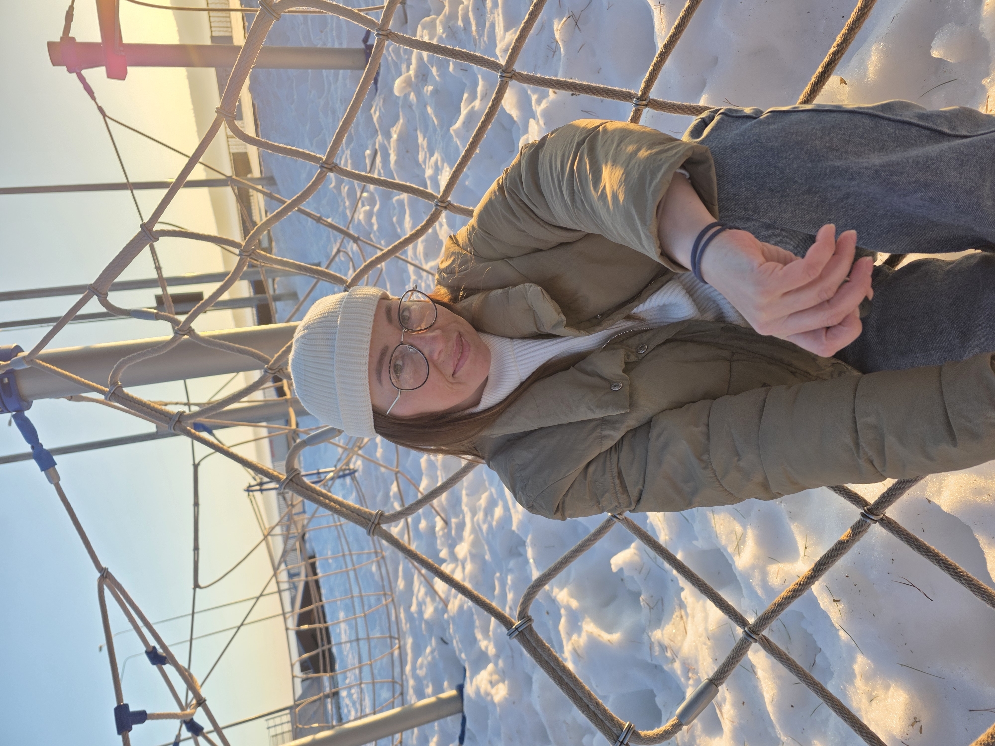 A medium shot shows a woman wearing a white beanie, glasses, a white sweater, and a beige puffy jacket, sitting in a rope net structure. She is facing the camera, smiling slightly. The background includes snow-covered ground and a metal structure with ropes. The lighting suggests it is daytime with soft sunlight. The photo was taken in the city of Chaikovsky, Russia.