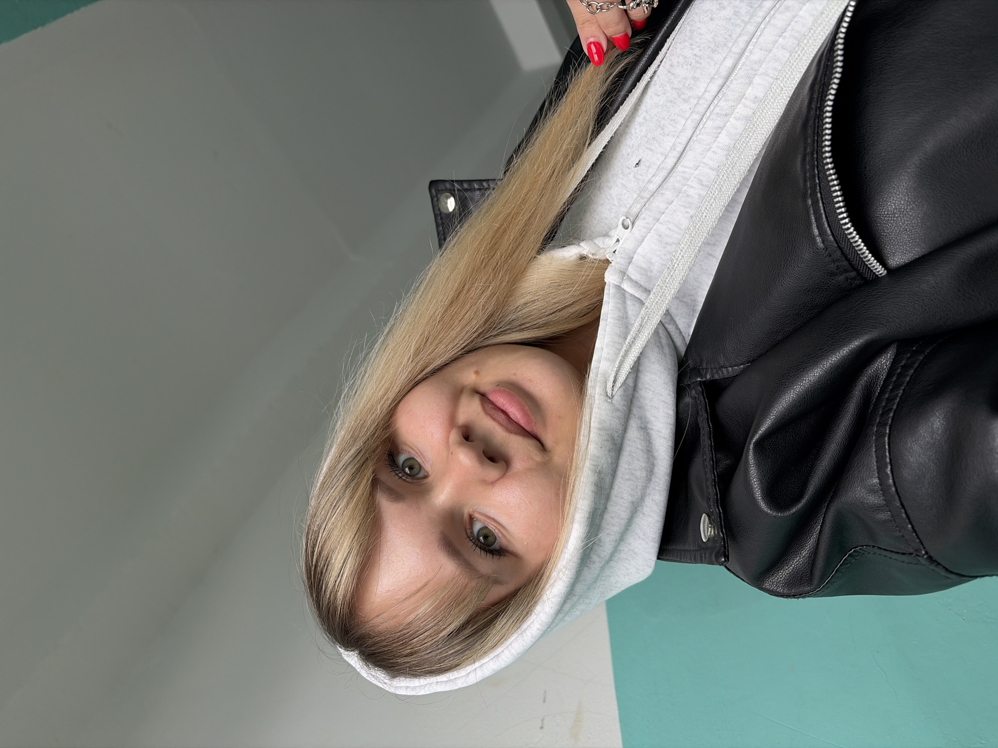 A young woman with blonde hair is posing for a selfie. She is wearing a gray hoodie and a black leather jacket. Her nails are painted red. The background is a two-tone wall, with gray and turquoise sections. The location appears to be indoors. There is no specific information to confirm the location in Chaikovsky, Russia.