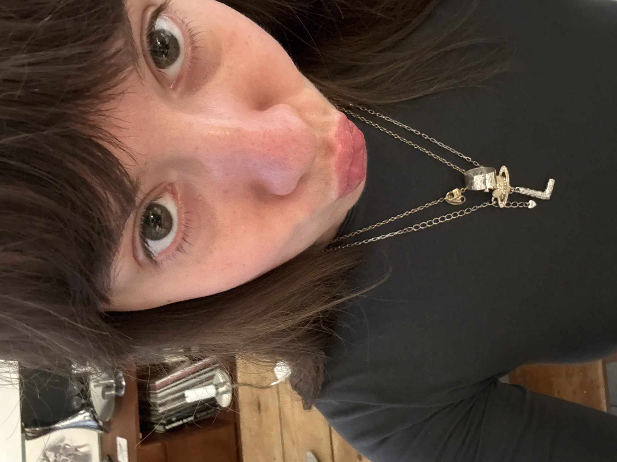 A close-up, tilted selfie captures a person in an indoor setting, likely in London, United Kingdom. The subject has fair skin, dark brown hair with bangs, and light-colored, possibly hazel or green, eyes. They are looking directly at the camera with a playful expression, their lips slightly puckered.
The person is wearing a plain, dark-colored long-sleeved top. A silver-toned chain necklace is prominent, adorned with several unique pendants. One pendant is a textured, hammered metallic ring. Another is a stylized figure, possibly a key or a letter (such as 'L' or 'J'), crafted from a lighter-toned metal and embellished with small, sparkling stones. A smaller charm, possibly heart-shaped, also hangs from the chain.
The background is softly blurred, revealing hints of a domestic environment. Visible elements include wooden surfaces, potentially floorboards or furniture, and stacked items that resemble vinyl records or books on a shelf. The lighting appears natural, suggesting daytime. The image conveys a casual and possibly humorous moment. No text is visible in the media.