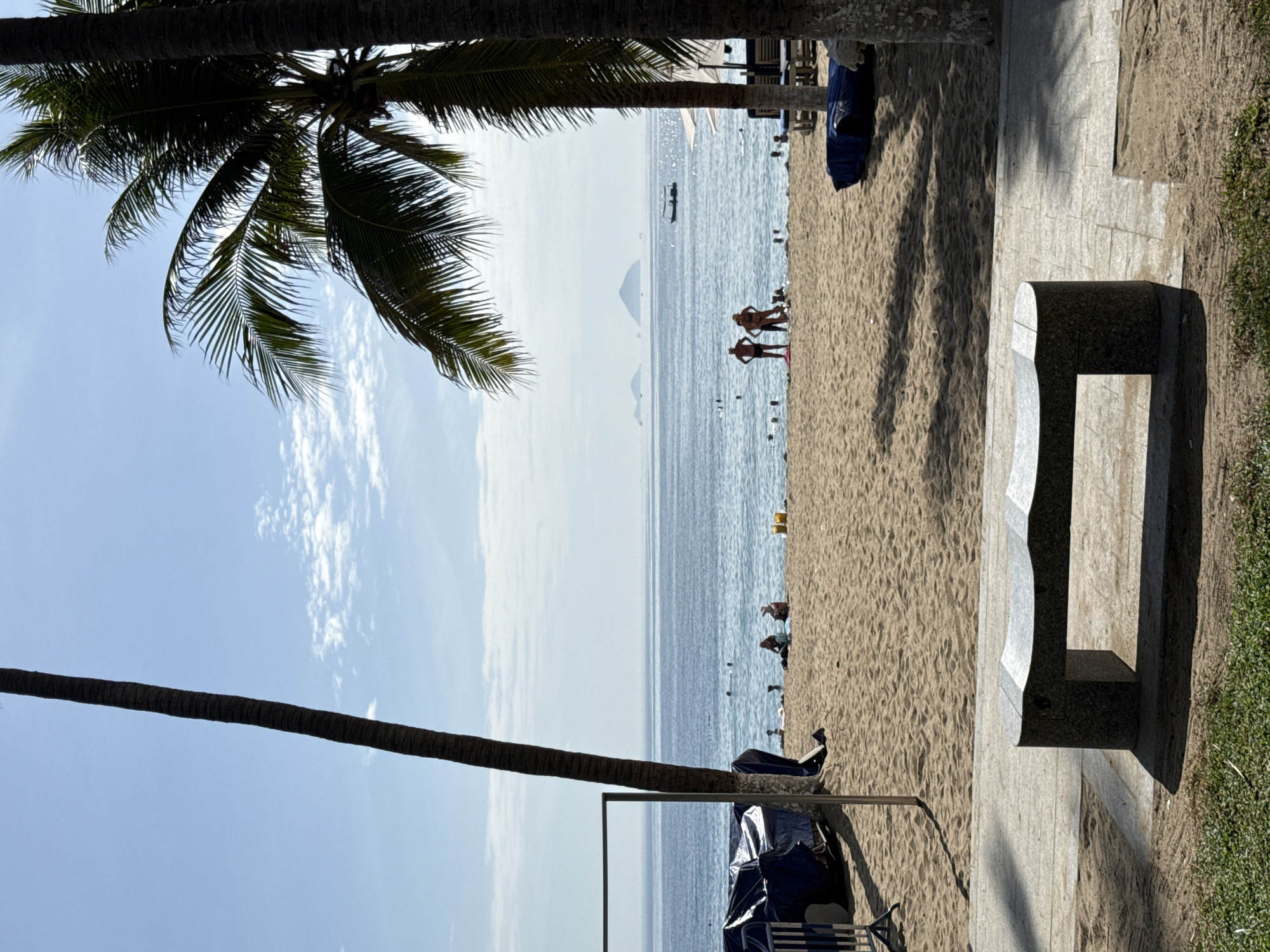 A long shot captures a sunny day at a beach in Nha Trang, Vietnam, with palm trees framing the scene. In the foreground, a modern, curved granite bench sits on a tiled pathway. Beyond the bench, a sandy beach stretches towards the ocean. Several people are visible on the beach, some sitting and relaxing, while others are wading in the shallow water or swimming further out. In the middle ground, a group of three individuals stands near the water's edge, silhouetted against the bright sea. The ocean itself is a calm blue, with a few small islands visible on the horizon. A small boat is also present on the water. The sky is a clear blue with scattered white clouds. The overall impression is one of a peaceful and vibrant beach day.