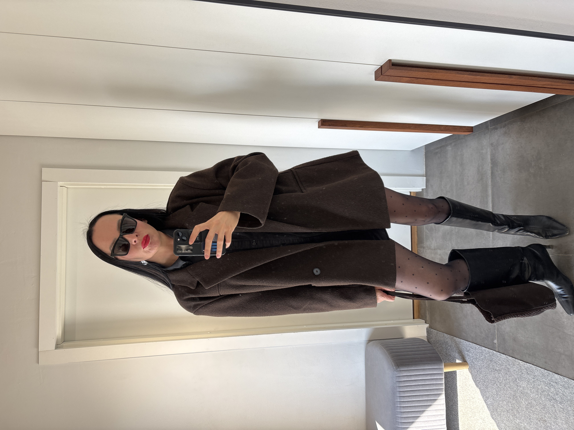 A full-body mirror selfie captures a woman standing indoors, dressed in a contemporary outfit. The woman has long, dark hair and is wearing black sunglasses, red lipstick, and has a neutral to slightly pouty expression. She is clad in a dark brown, long coat or jacket over a black top or dress, paired with sheer black stockings featuring small polka dots, and black knee-high boots with heels. She holds a smartphone in her right hand, positioned to take the photo, while a dark brown handbag hangs from her left arm.
The setting is an interior space, possibly a hallway or dressing room, characterized by bright, even lighting suggesting daytime. The background includes white walls and a white ceiling adorned with horizontal wooden beams or decorative slats. The floor is covered in large grey tiles, and a light grey upholstered bench with wooden legs rests on a grey rug or carpet on the right side of the frame. No visible text is present in the image. The location context is Chaikovsky, Russia.