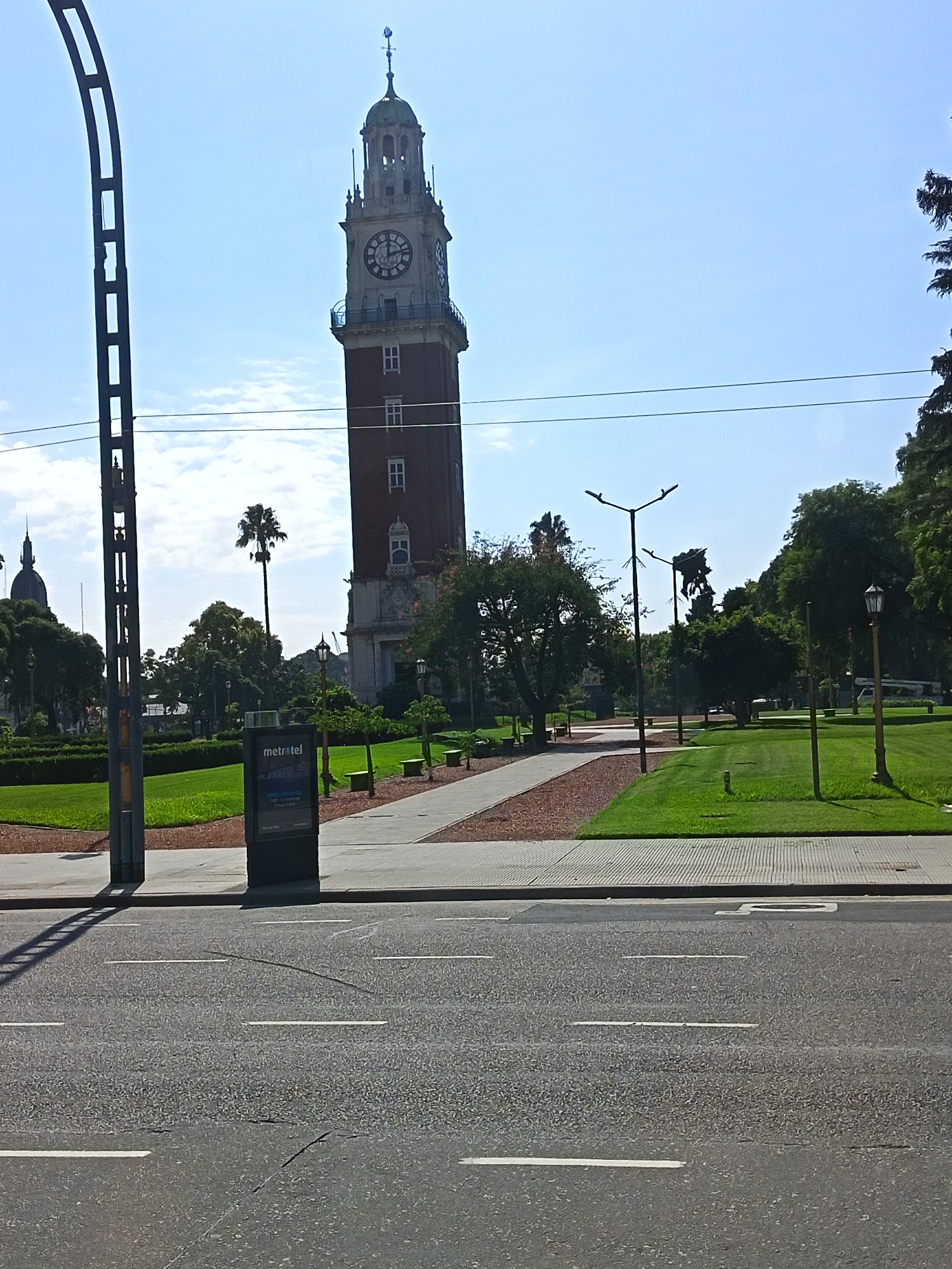 The image shows a daytime view of the Torre Monumental (also known as the Torre de los Ingleses or English Tower) in Buenos Aires, Argentina. The tower, a tall brick structure with a clock face, dominates the background. In front of the tower is a park with green lawns, trees, and walkways. Streetlights line the walkway. A Metrotel advertising sign is visible on the edge of the park near the sidewalk. In the foreground is a paved road with dashed white lines. A metal archway is located on the left side of the frame. No people are immediately visible in the scene. The sky is light blue with some clouds.