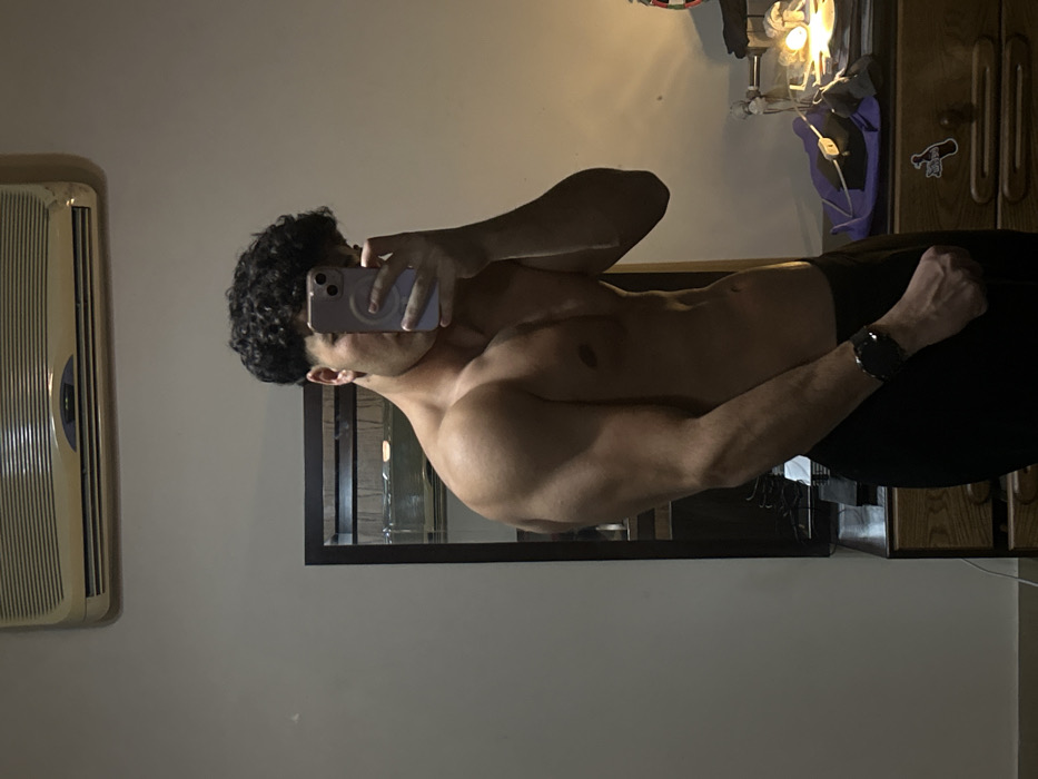 A shirtless man with curly hair stands sideways, holding a phone up to obscure part of his face. He wears black pants and a wristwatch. In the background, a window air conditioning unit is on the left, while a dark framed mirror hangs on the wall behind him. A wooden dresser, a hanging jacket, and a lamp are also visible.