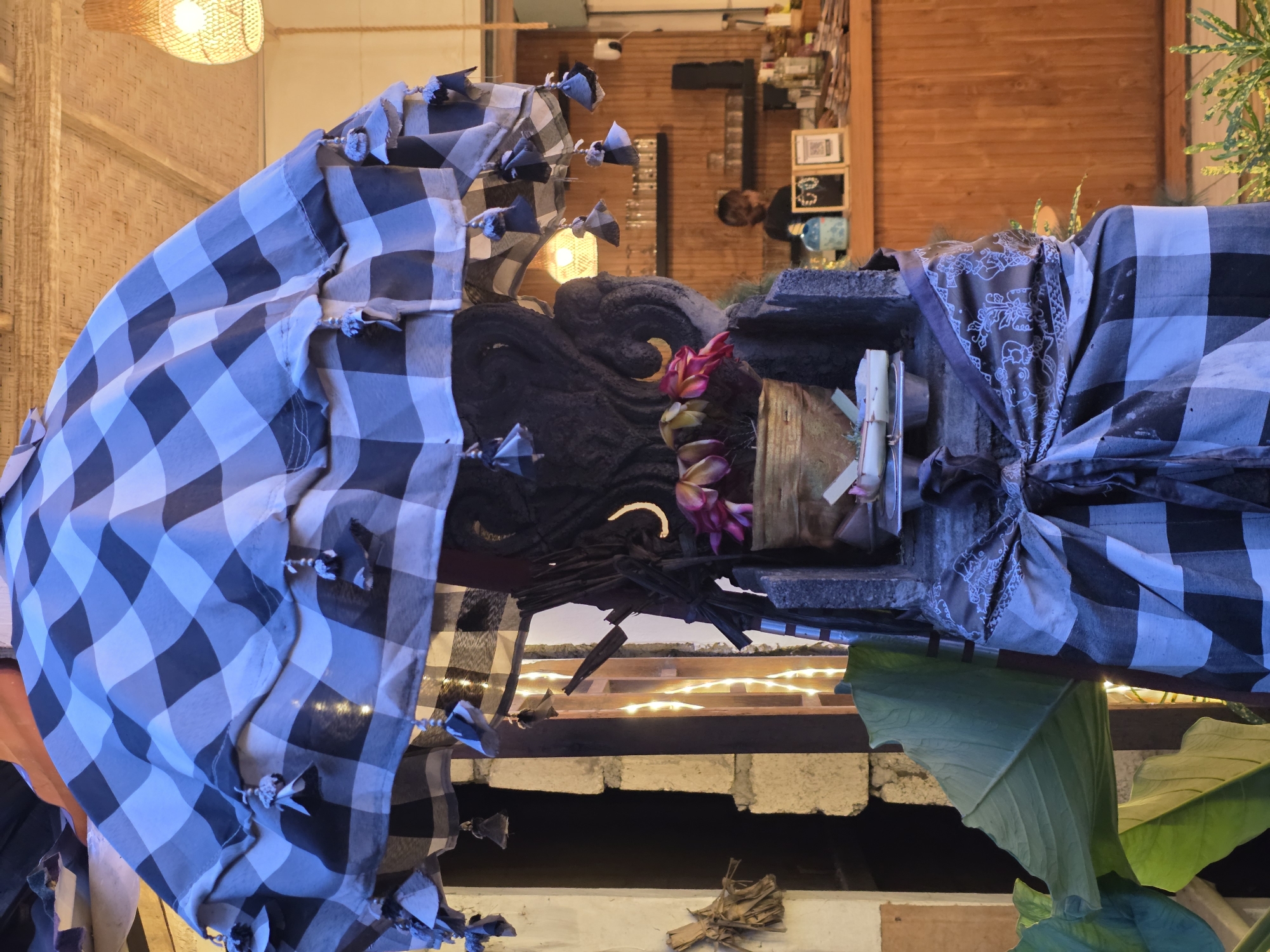 Here is a detailed description of the image:
The image captures a close-up view of a culturally significant display. The focal point is a statue or effigy adorned with a blue and white checkered fabric, styled as if it were draped clothing. There is dark, textured material which forms the head or upper body of the statue, decorated with flowers, possibly plumeria, adding a touch of color.
The statue is placed on a ledge, with warm white fairy lights visible underneath, providing a soft, ambient glow. A large green leaf from a tropical plant extends into the frame in the lower right corner, suggesting a lush or natural setting.
The background offers hints of an interior space. A woven bamboo-like panel is on the left, with a warm yellow light fixture above it. Directly behind the statue, a glimpse of the room's interior shows wooden walls, a man standing in the background, and a couple of chalkboards or displays. This suggests the effigy is placed in a restaurant, maybe in Bali.
The overall ambiance is that of a place with a Balinese cultural atmosphere. The presence of the statue, its decorations, and the choice of decor all point to this cultural identity.