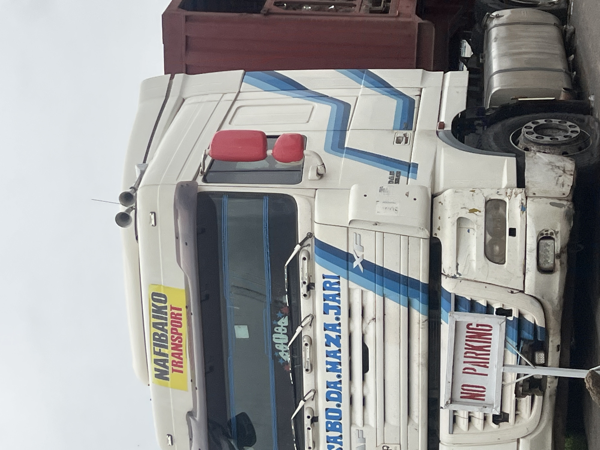 This is a medium shot, eye-level, outdoor photograph of a white DAF transport truck parked on an unpaved area, possibly a road or a loading bay, in Jalingo, Nigeria. The truck has a red trailer attached and features blue and white stripes along its side. A yellow sticker with the words "NAFIBAIKO TRANSPORT" is visible on the top of the cabin. A red sign reading "NO PARKING" is mounted on a pole in front of the truck. The truck's front tire on the left side appears deflated or damaged. The sky is overcast and a large fuel tank is visible on the right side of the truck. The overall scene suggests the truck is either temporarily out of service or is awaiting transport or parking instructions.