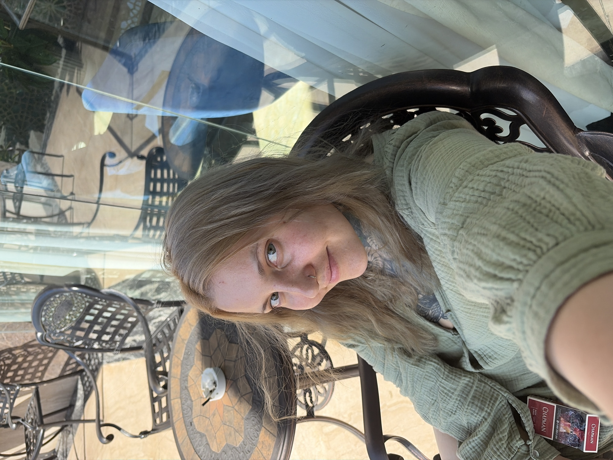 A fair-skinned individual with light brown hair sits in a dark brown metal chair. A green textured top is being worn. A pack of "Charman" brand cigarettes is visible. The location appears to be an outdoor patio or garden area, with additional metal chairs and a table with a mosaic tile top. Reflected in a glass barrier are views of additional outdoor furniture and blue umbrellas, suggesting a restaurant or café setting. It is not possible to verify the location as Đà Nẵng, Vietnam, without additional contextual information.